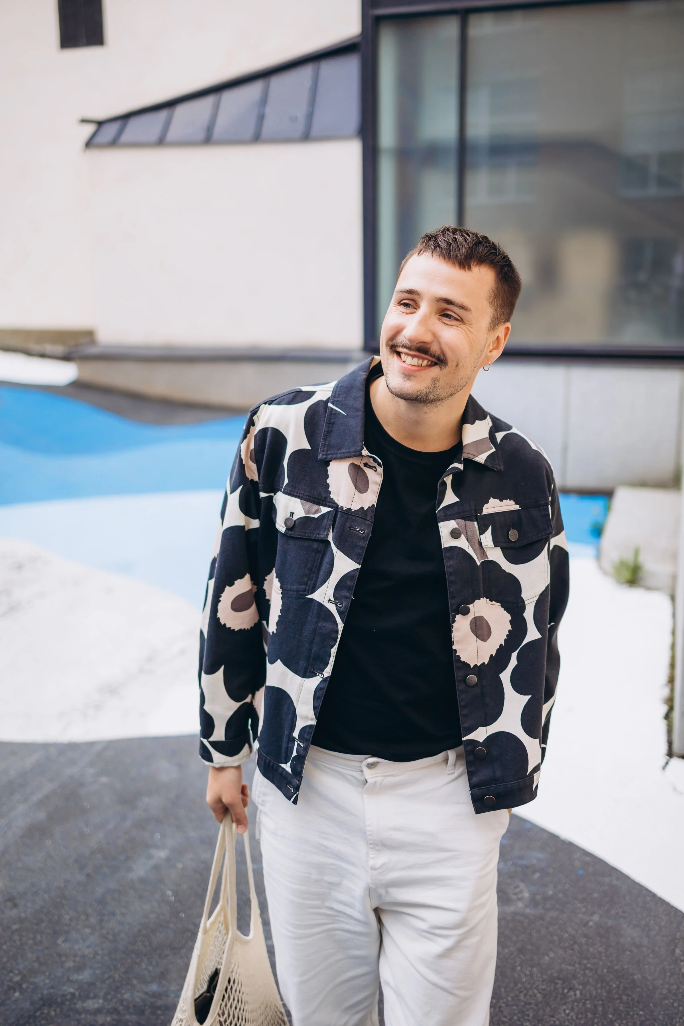 Pesojoonas hymyilee kädessä kangaskassi, taustalla moderni rakennus ja graffitiseinämä.