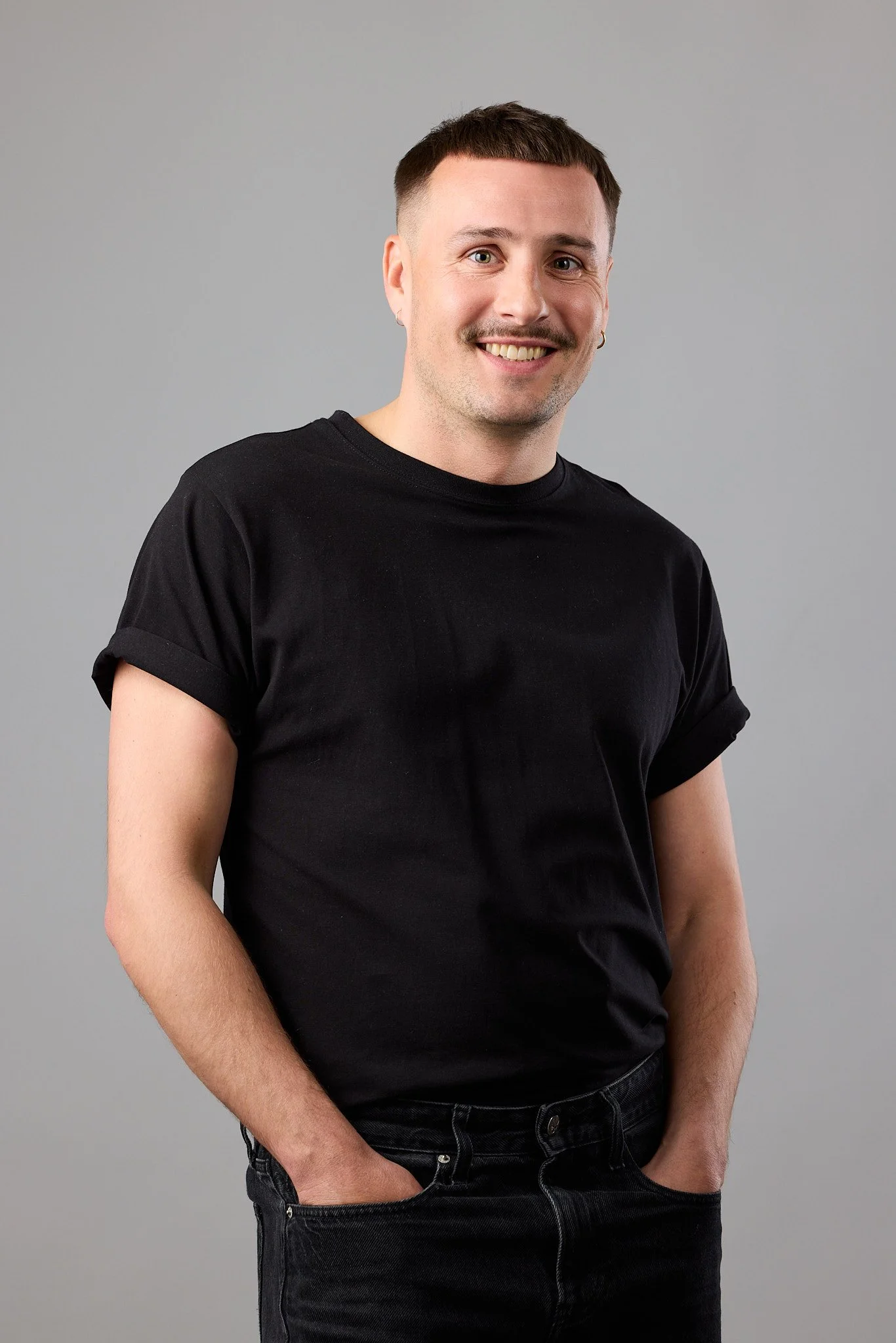 Pesojoonas hymyilee ja katsoo kameran suuntaan, pukeutuneena mustaan t-paitaan ja farkkuihin, taustalla vaalea harmaa.