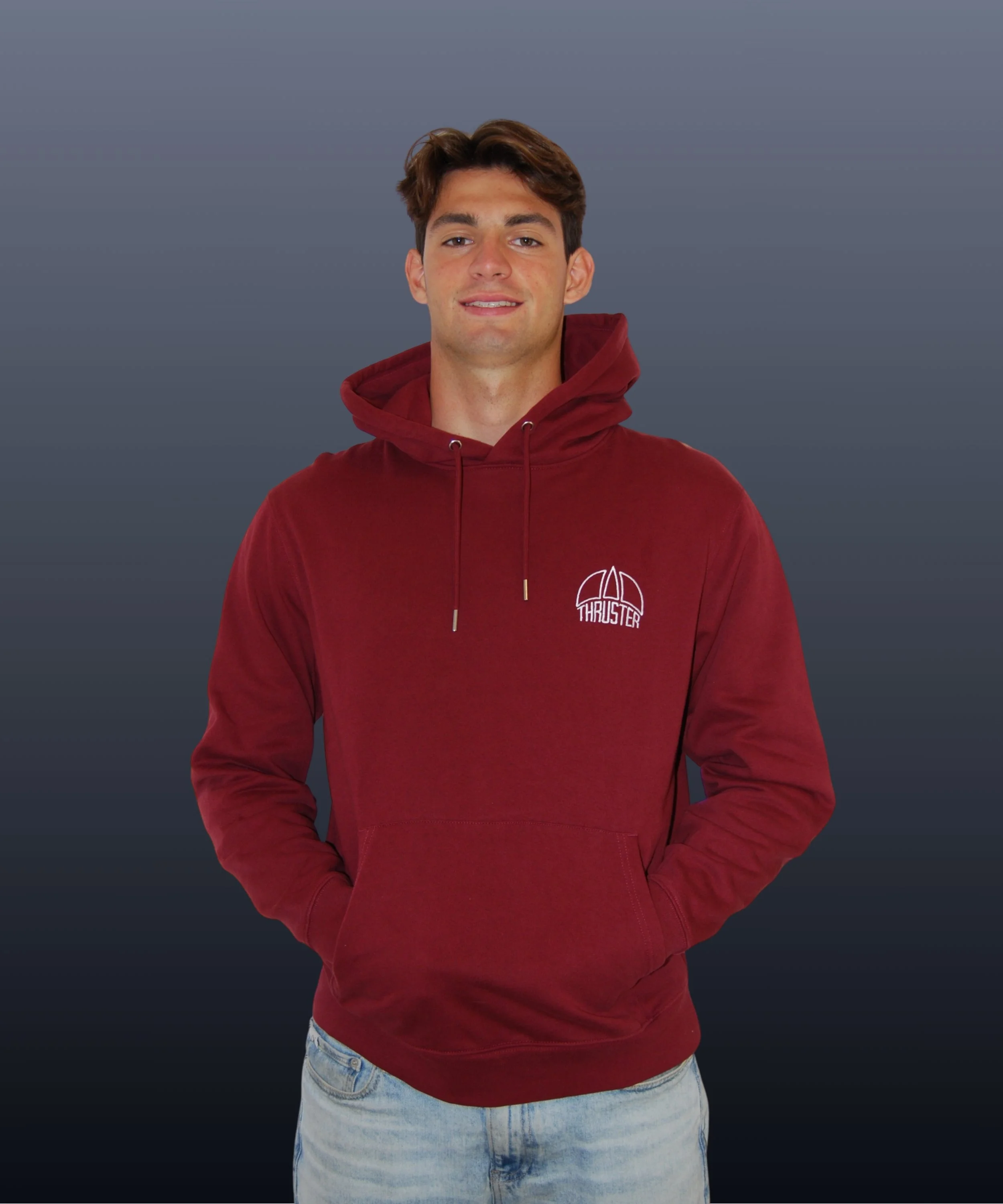 Giovane uomo con capelli castani, indossa una felpa rossa con cappuccio e pantaloni di jeans chiari, sfondo grigio scuro.