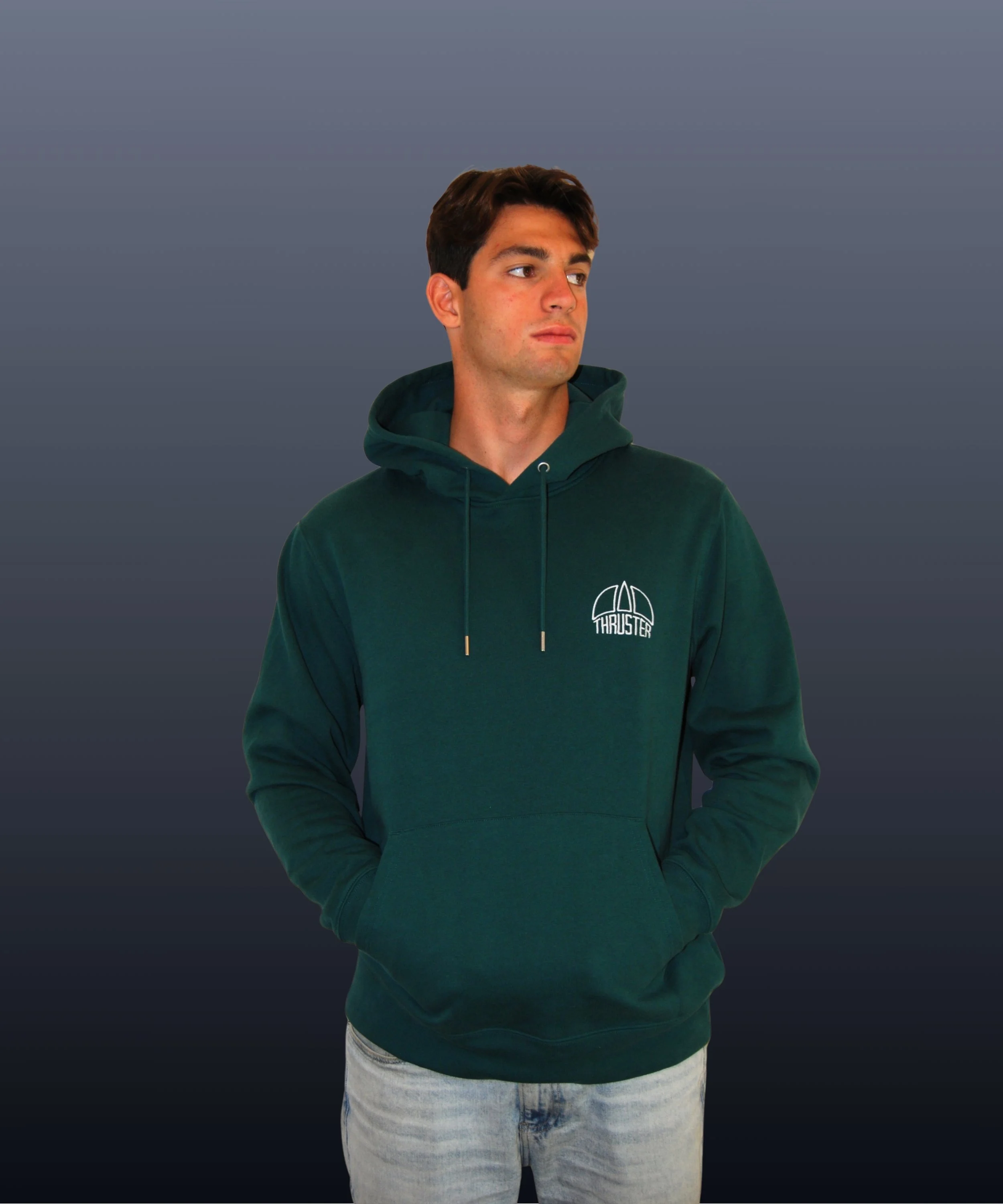 Giovane uomo con giacca felpata verde scuro con cappuccio, logo THRUSTER, in piedi contro sfondo sfumato grigio, ha i capelli castani, viso neutro e mani in tasca.