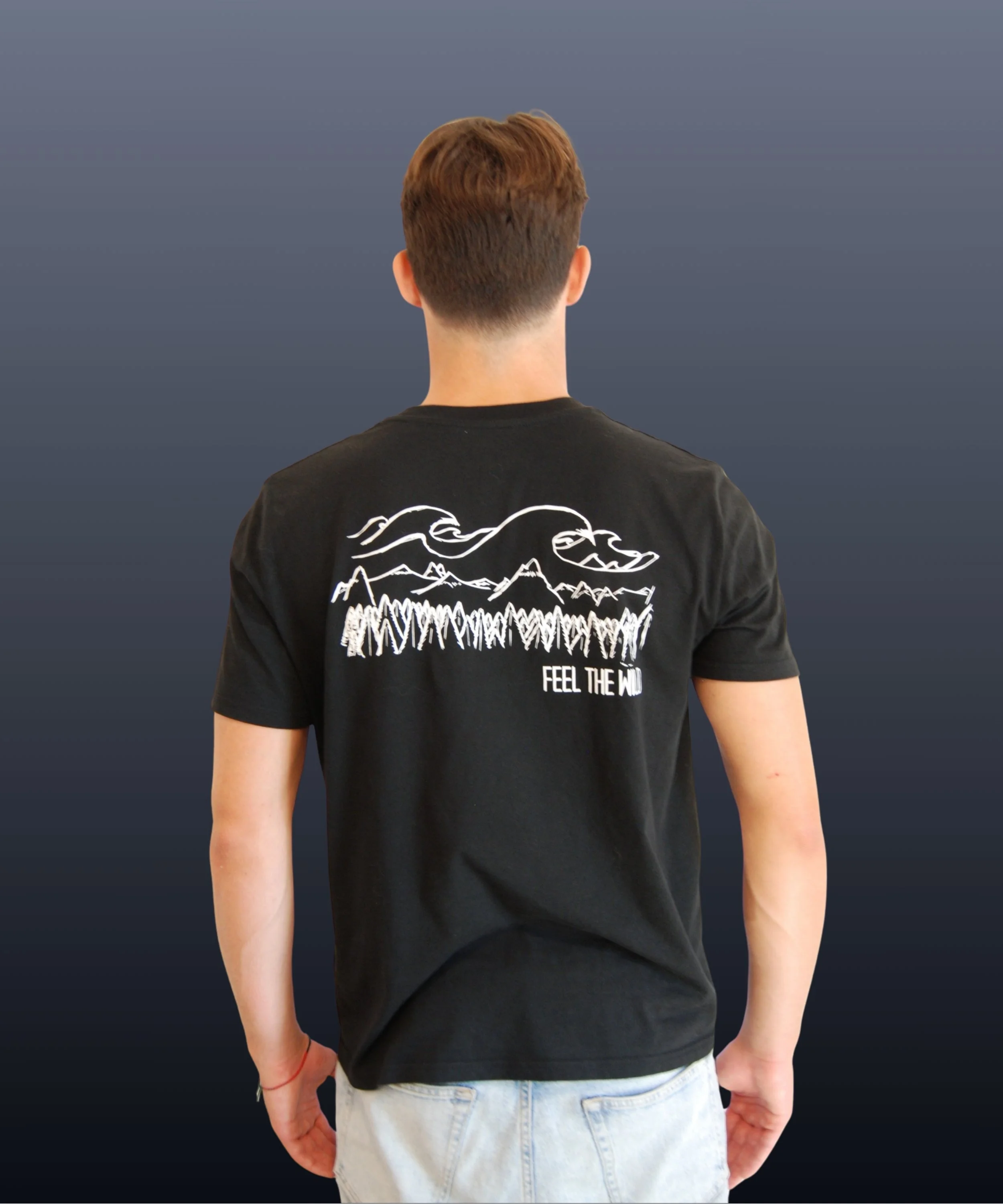 Retro di un uomo visto da dietro, indossa una maglietta nera con un disegno di montagne, onde e alberi, con la scritta 'FEEL THE WILD'