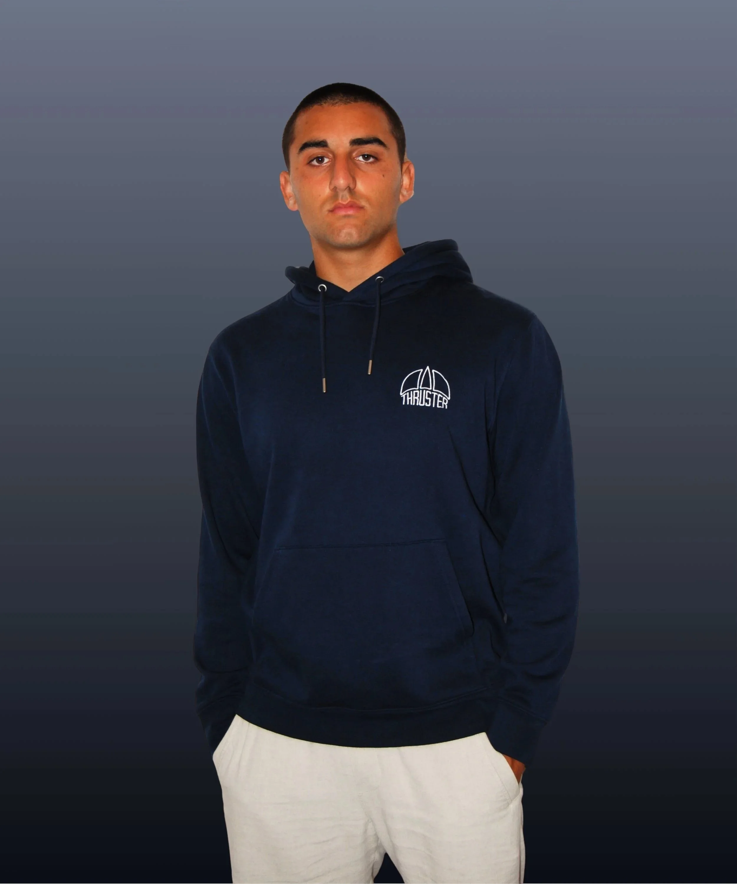 Ragazzo con cappuccio blu scuro con logo "Thruster" sulla maglia, posa con mani in tasca, sfondo grigio