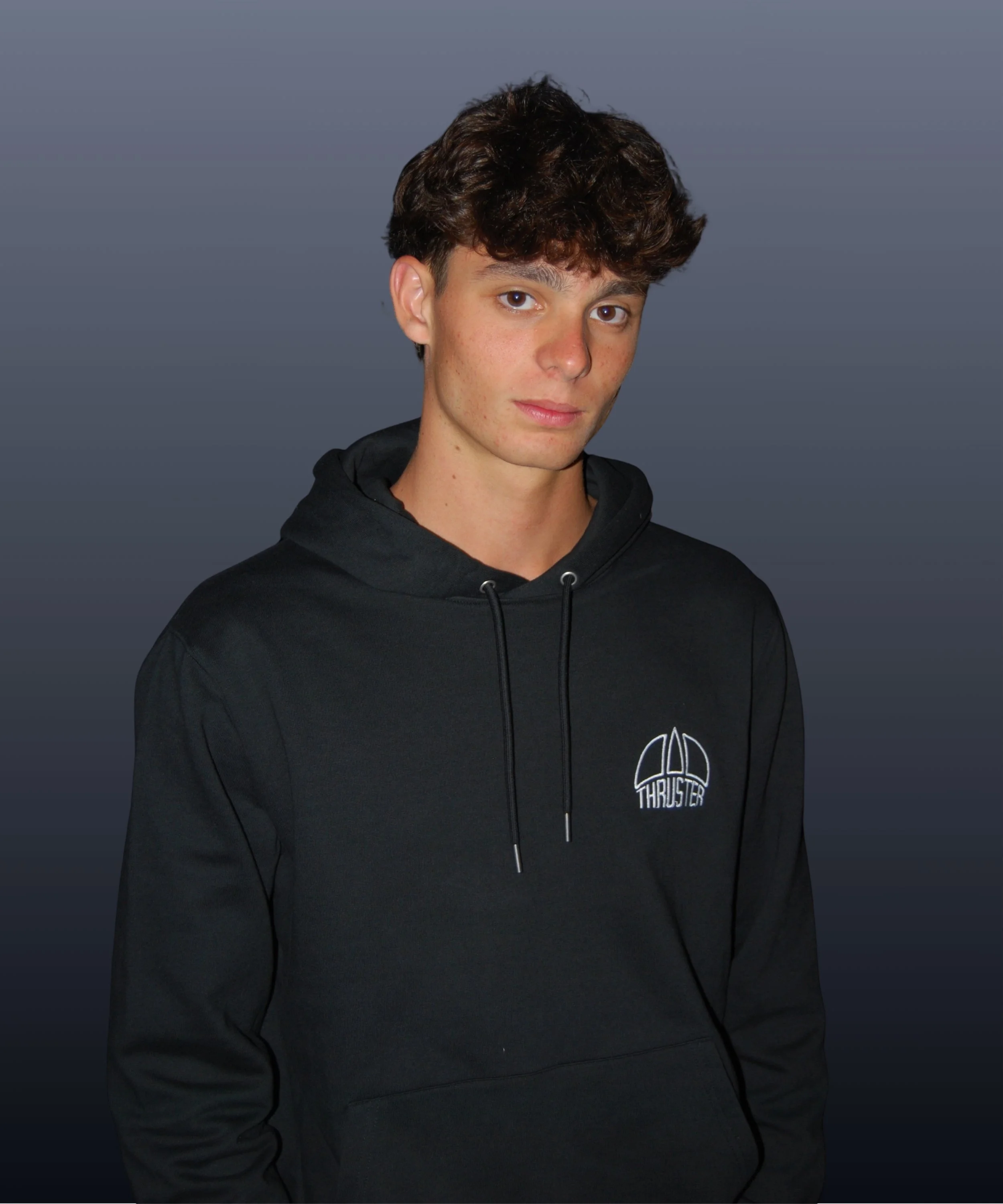 Ragazzo con capelli castani corti, indossa felpa nera con cappuccio e logo 'THRUSTER' sul petto, sfondo grigio scuro.