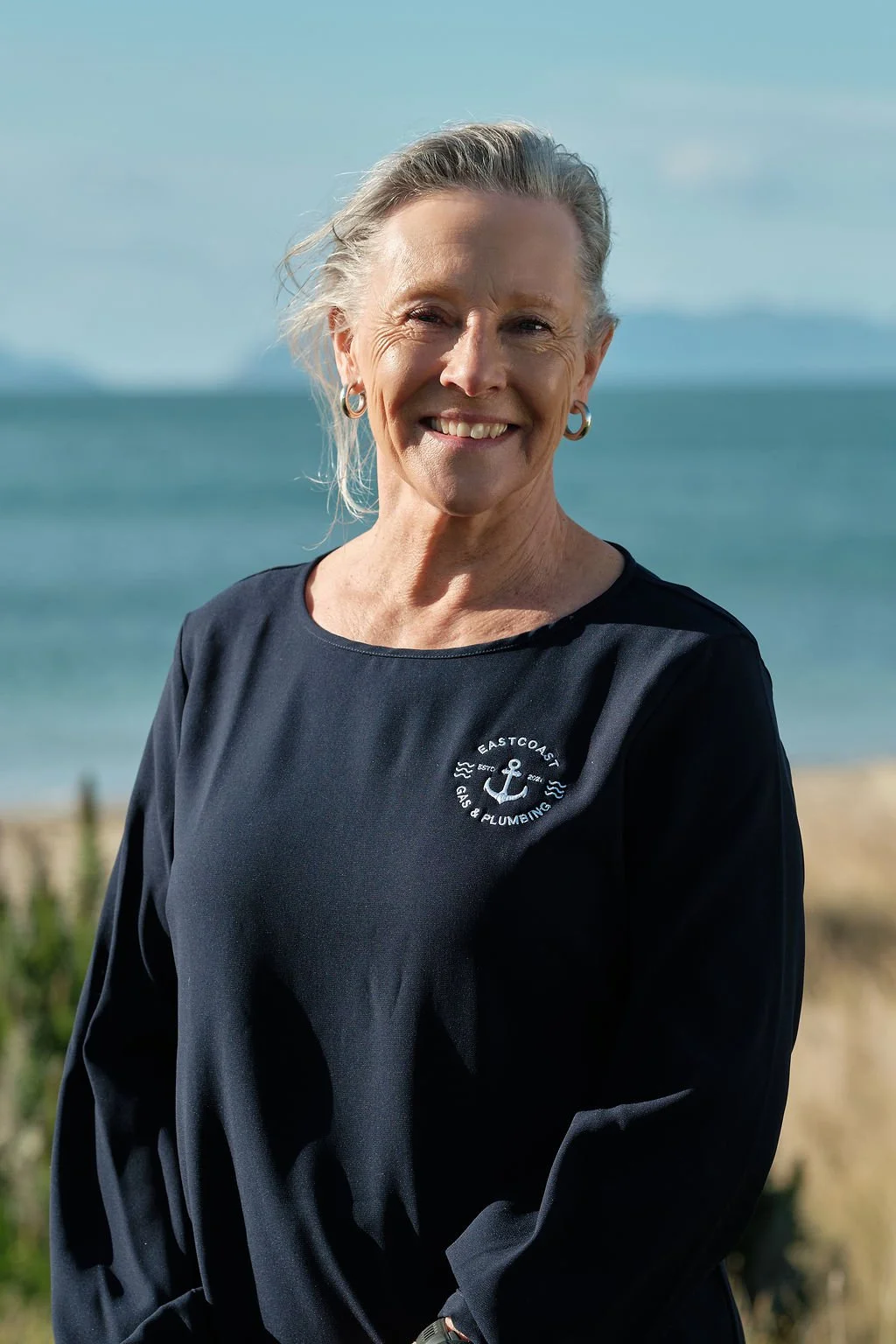 A woman wearing a black shirt with a logo that says 'East Coast Gas & Plumbing' with an anchor emblem, standing outdoors near the beach with ocean waves in the background and a cloudy sky.