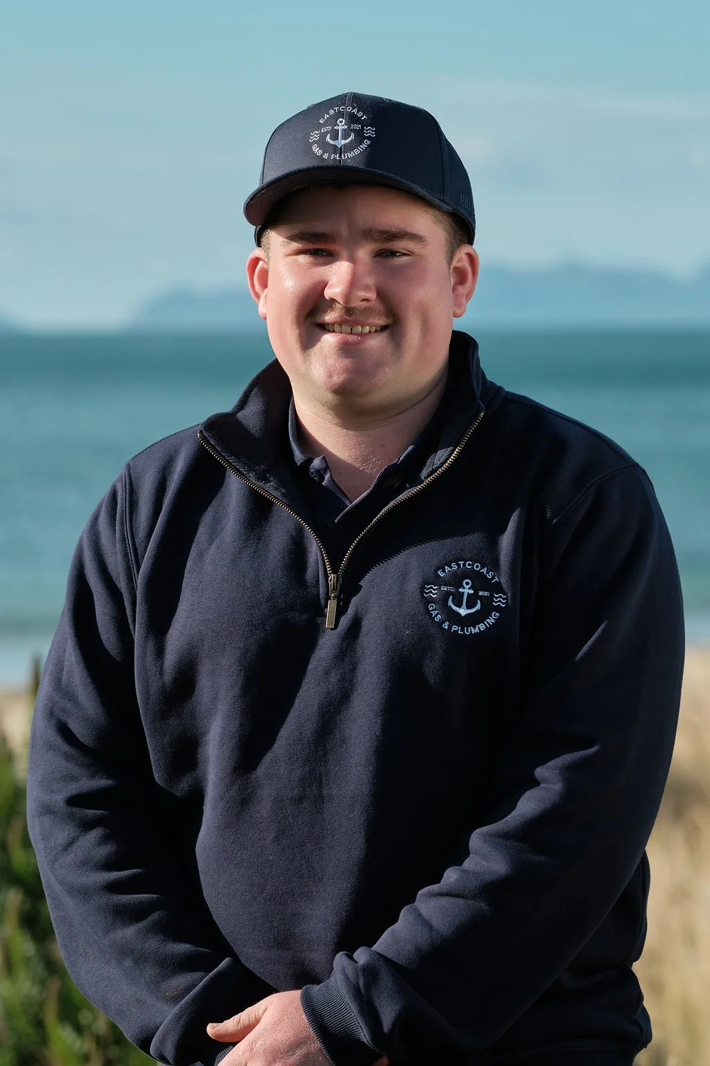 A man wearing a black cap and jacket with 'Eastcoast Gas & Plumbing' logo, standing outdoors near the ocean with a cloudy sky in the background.