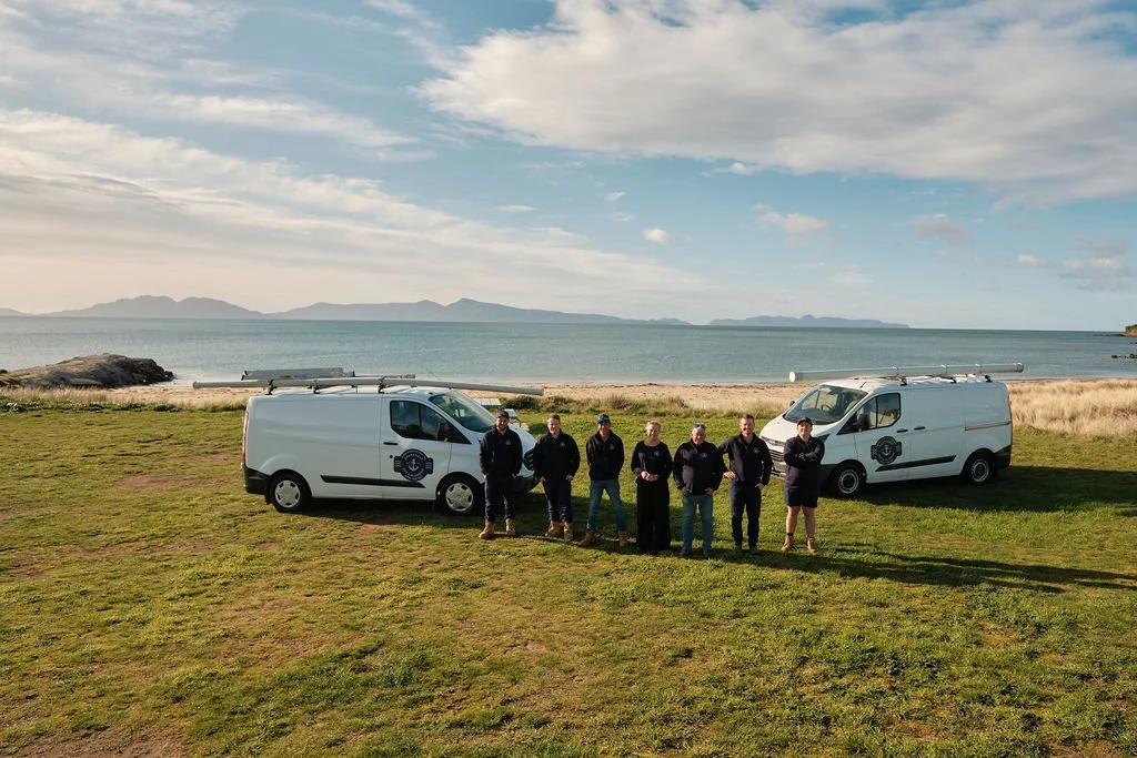 Group of seven people standing in front of two white service vans with surfboards on top, on a grassy area near a beach with water and distant hills in the background.
