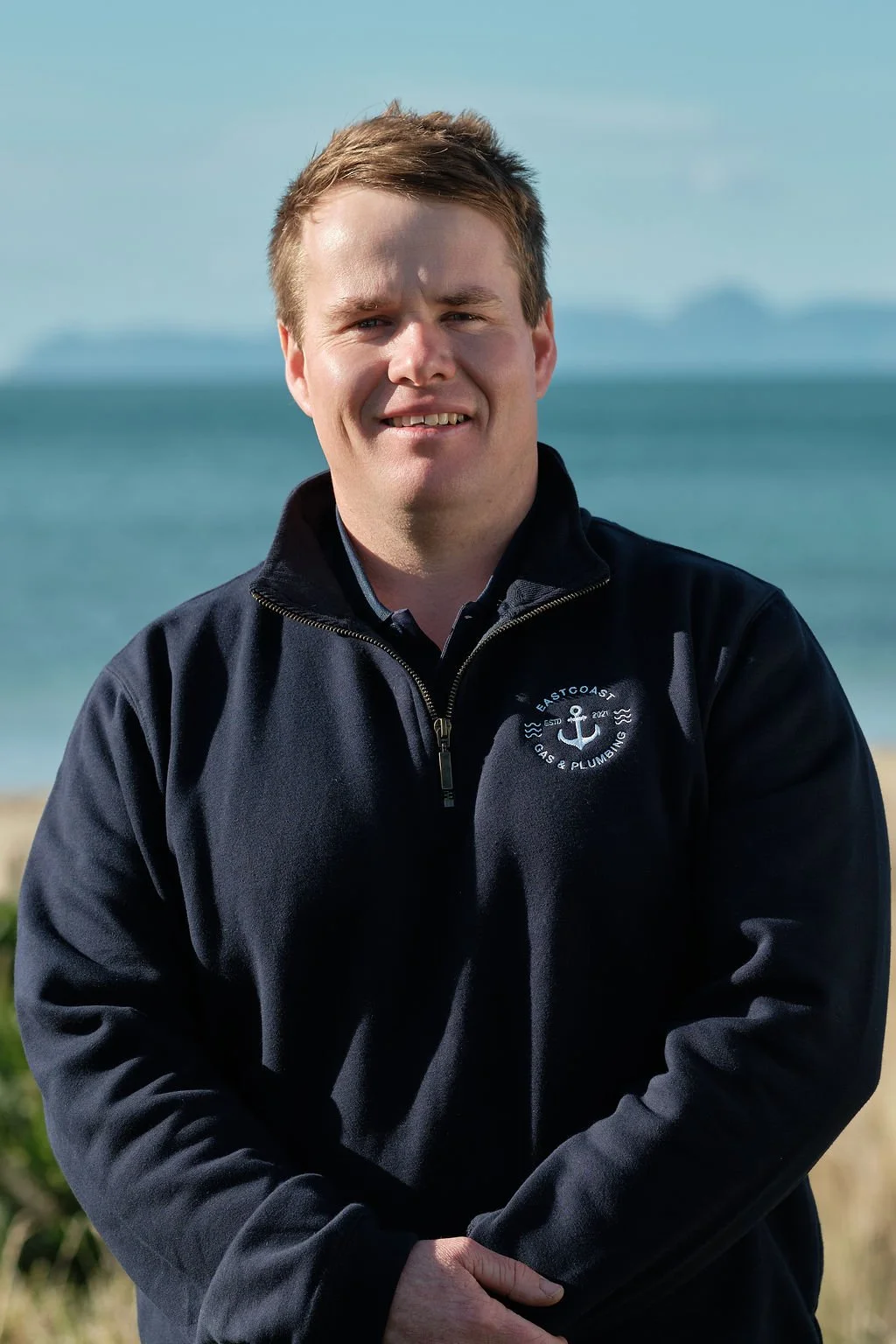 A man standing outdoors near a beach with the ocean in the background, wearing a black jacket with an anchor logo and the text 'East Coast Gas & Plumbing'.