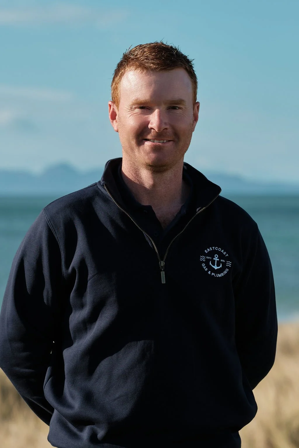 A man with short hair smiling outdoors, dressed in a black jacket with a logo on the chest that reads 'East Coast Gas & Plumbing'.