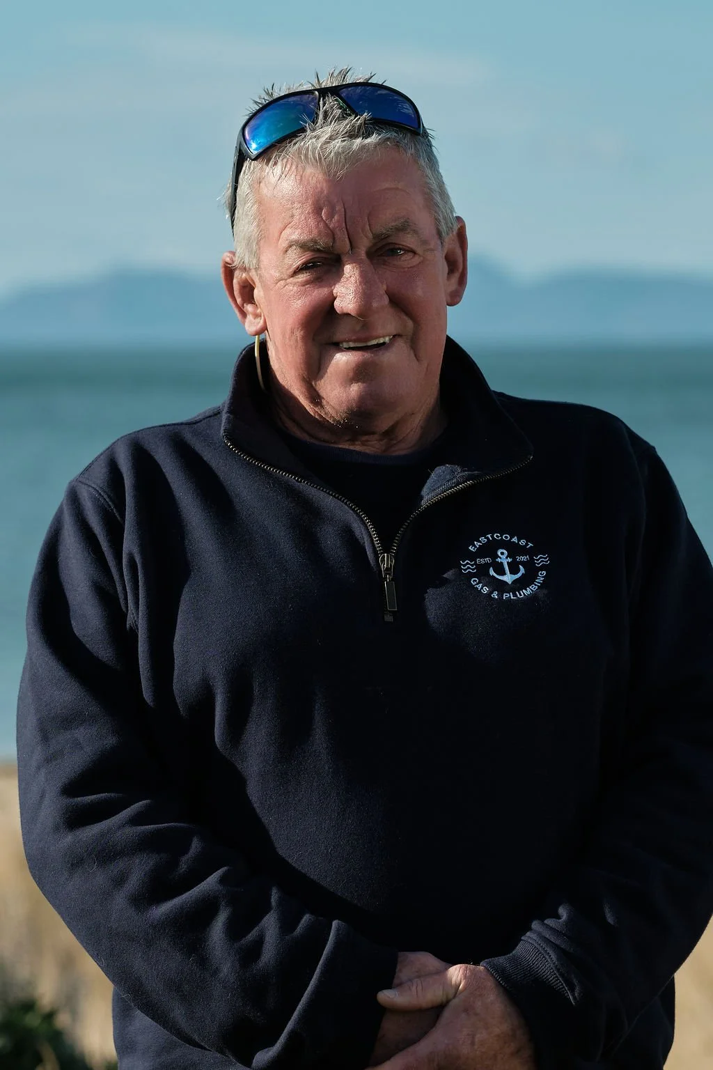 A man with gray hair and a black jacket with a logo, standing outdoors near the water with mountains in the background.