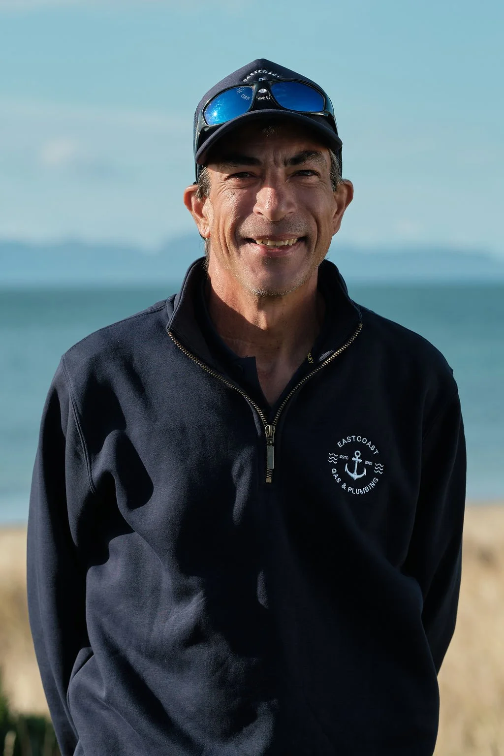 A man smiling outdoors near the water, wearing a black zipped jacket with an anchor logo and sunglasses on his cap.