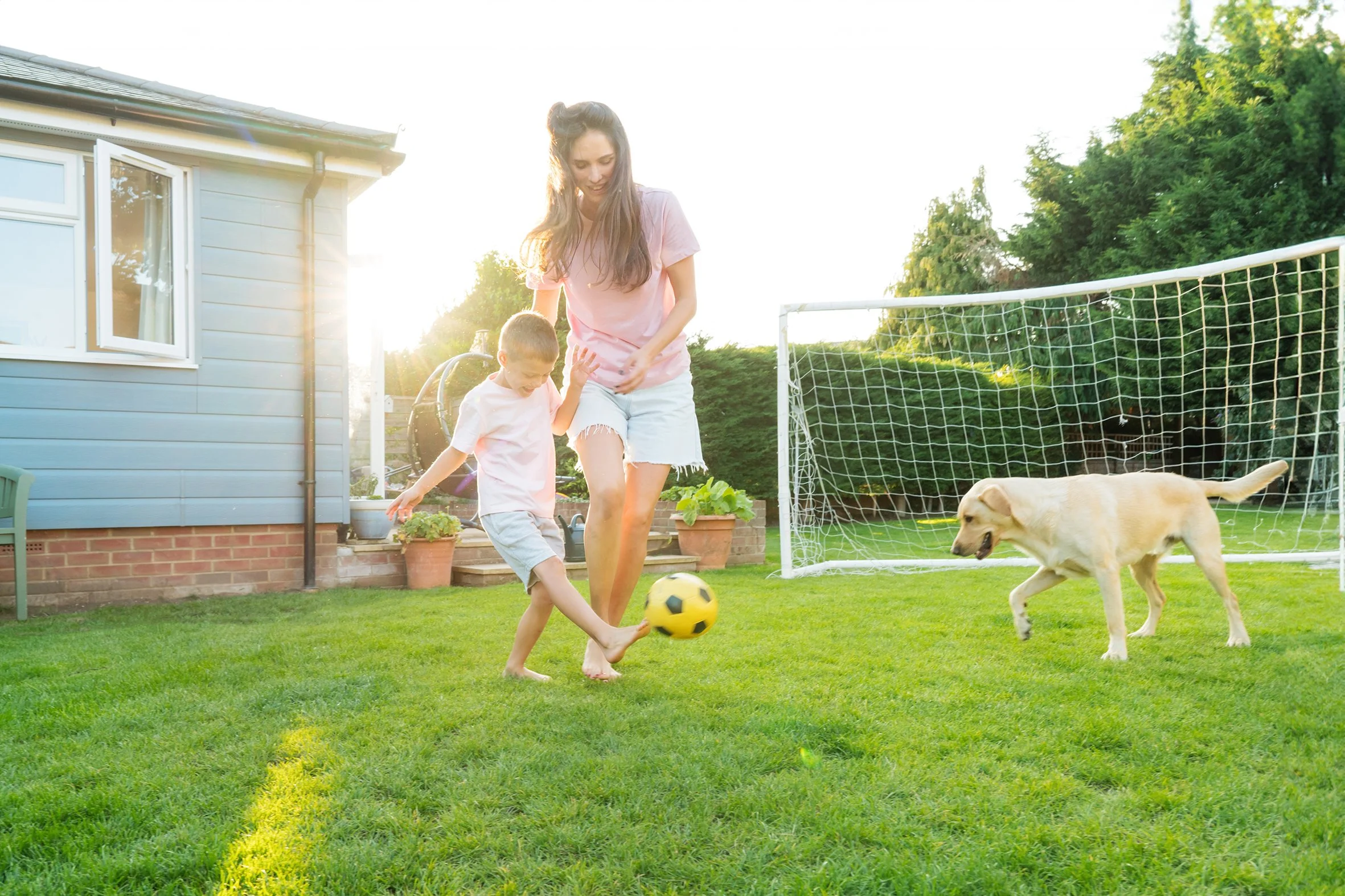 A woman and young boy playing soccer with a black and yellow ball on a green lawn with a dog nearby and a soccer goal in the background, during sunset.