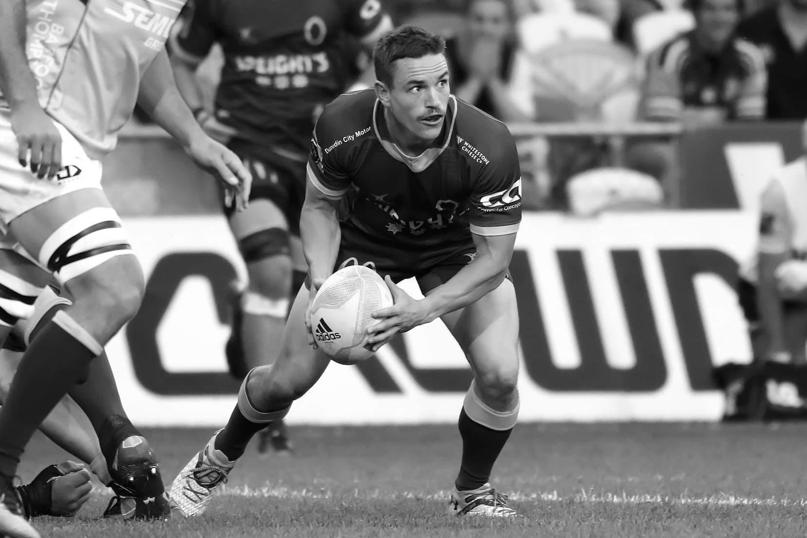 A rugby player in black uniform holding a rugby ball, preparing for a play during a match, with other players and a blurred crowd in the background.