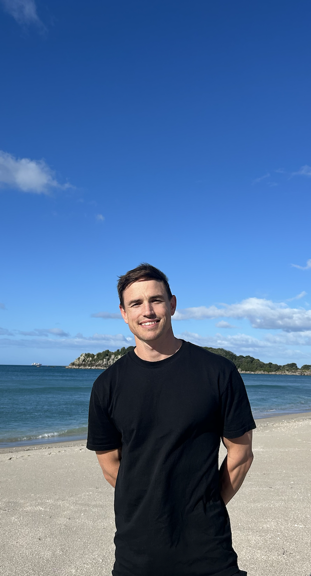 A young man with short dark hair, wearing a black t-shirt, standing on a sandy beach with clear blue water and an island in the background under a partly cloudy sky.