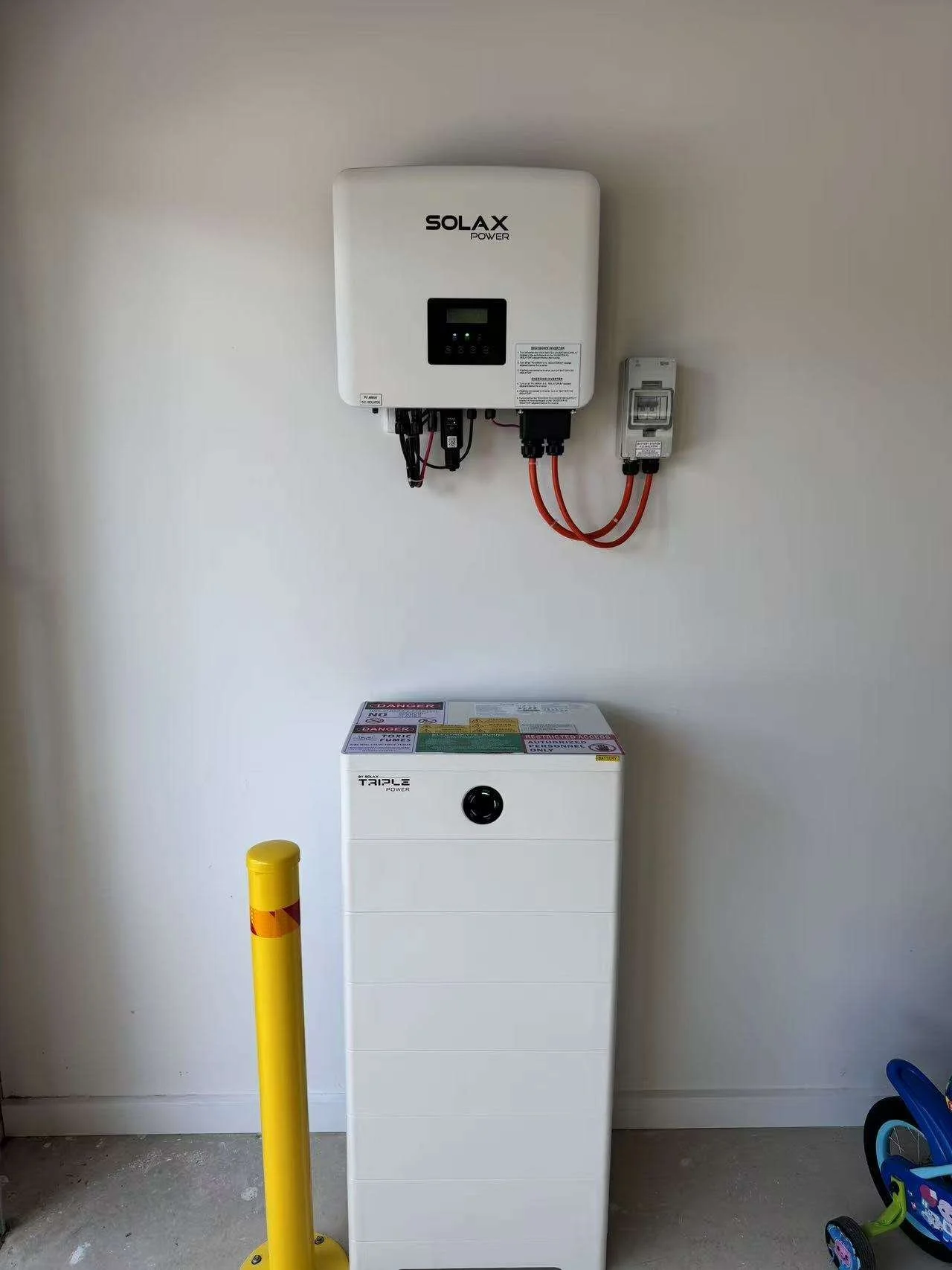 An electrical panel and a solar inverter mounted on a white wall, with a white battery storage unit below, and a yellow safety bollard in front.