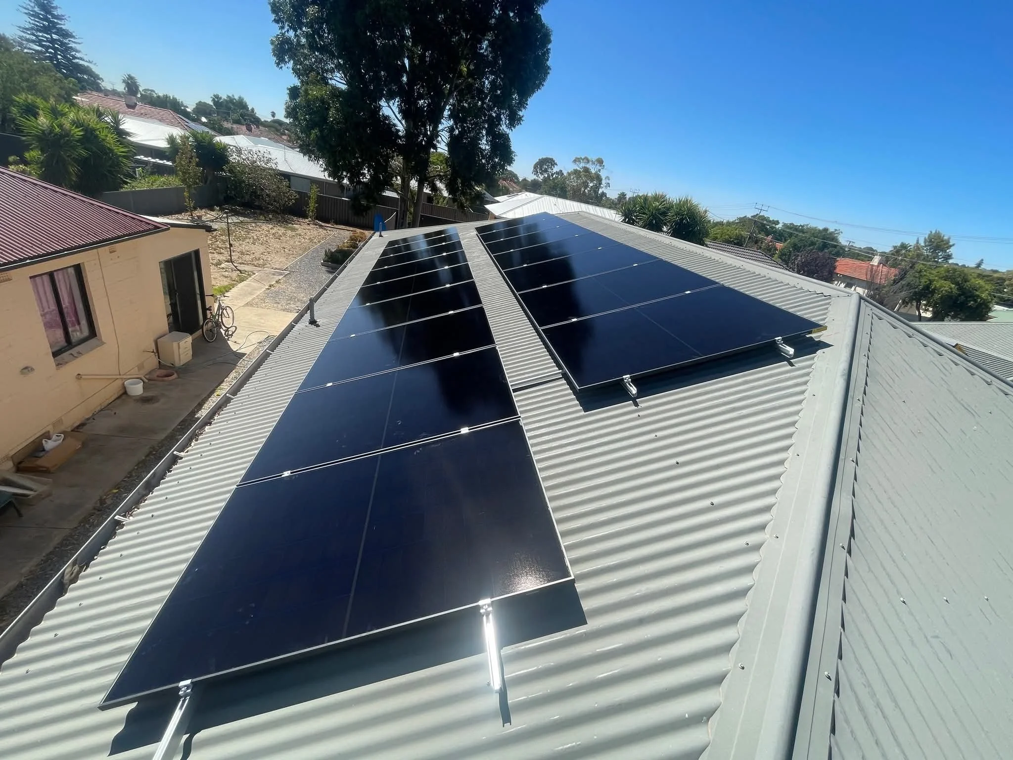 Solar panels installed on a corrugated metal roof of a house on a sunny day, with trees and neighboring houses in the background.