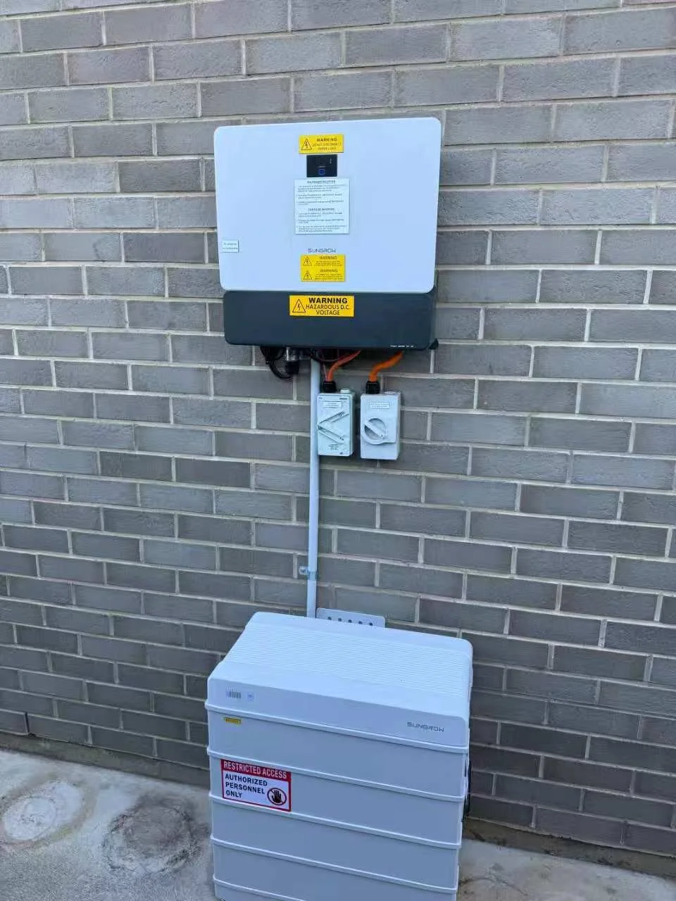 Photo of a solar power inverter and storage system installed on a brick wall outdoors. The inverter has warning labels, and the storage unit has a