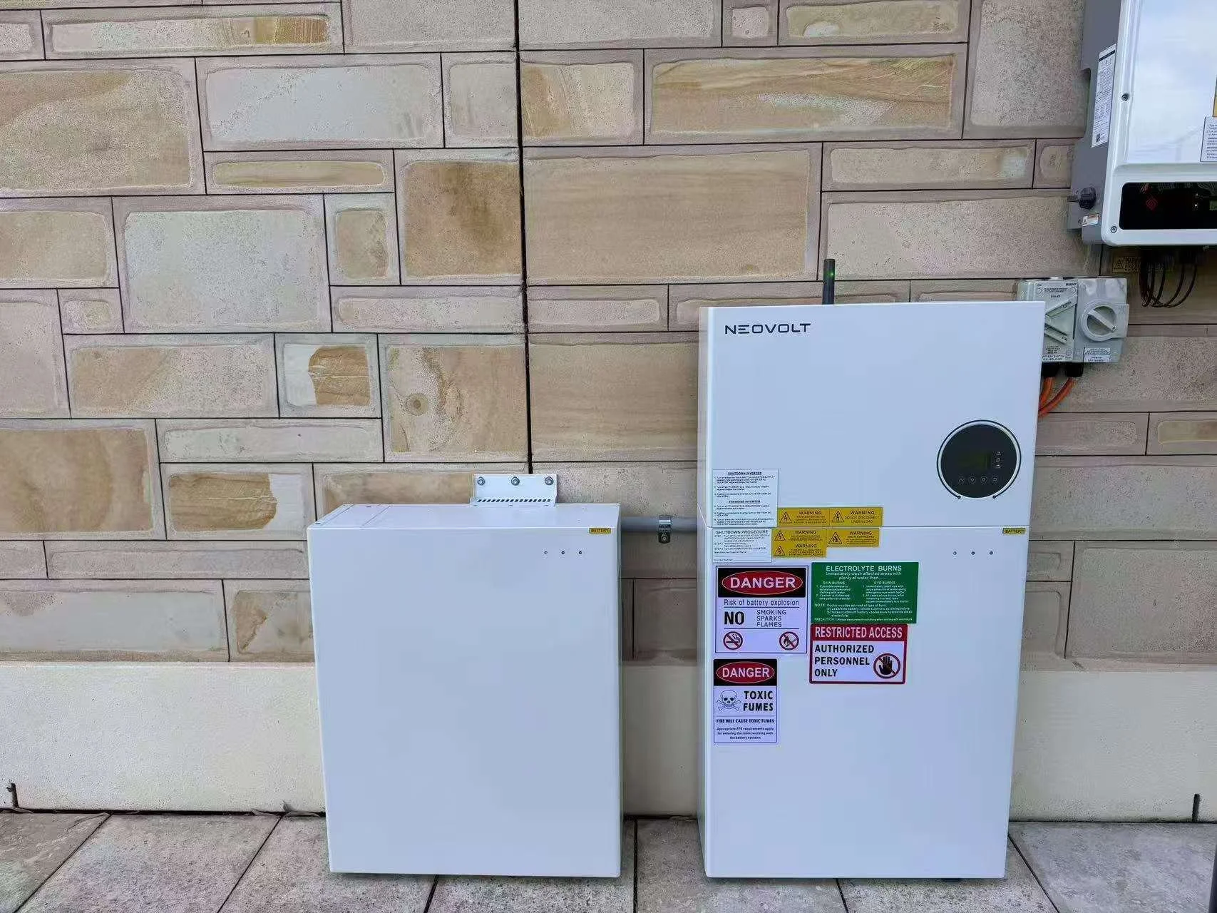 Two white electrical boxes mounted on a beige brick wall, with warning and safety labels on the larger box.