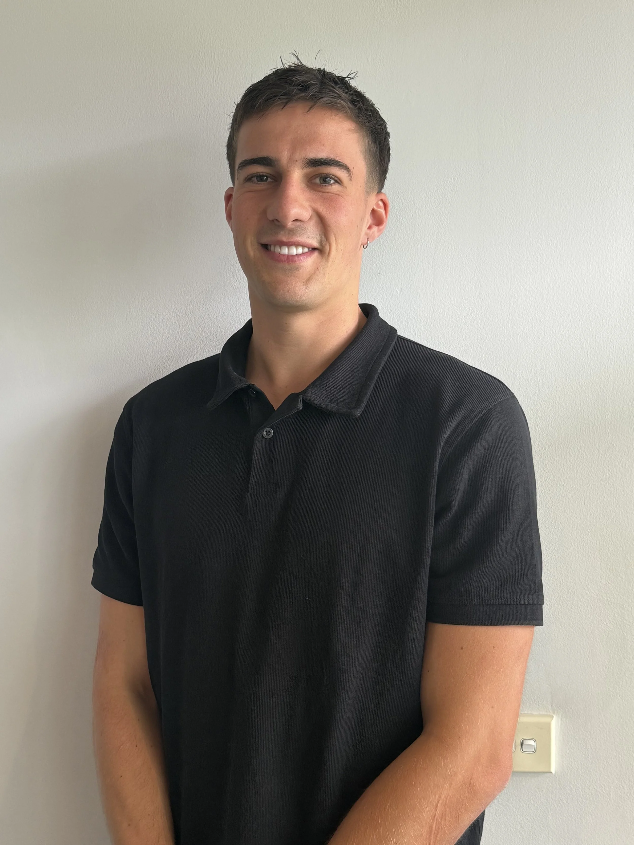 A young man with short dark hair, wearing a black polo shirt, smiling and standing against a plain white wall.