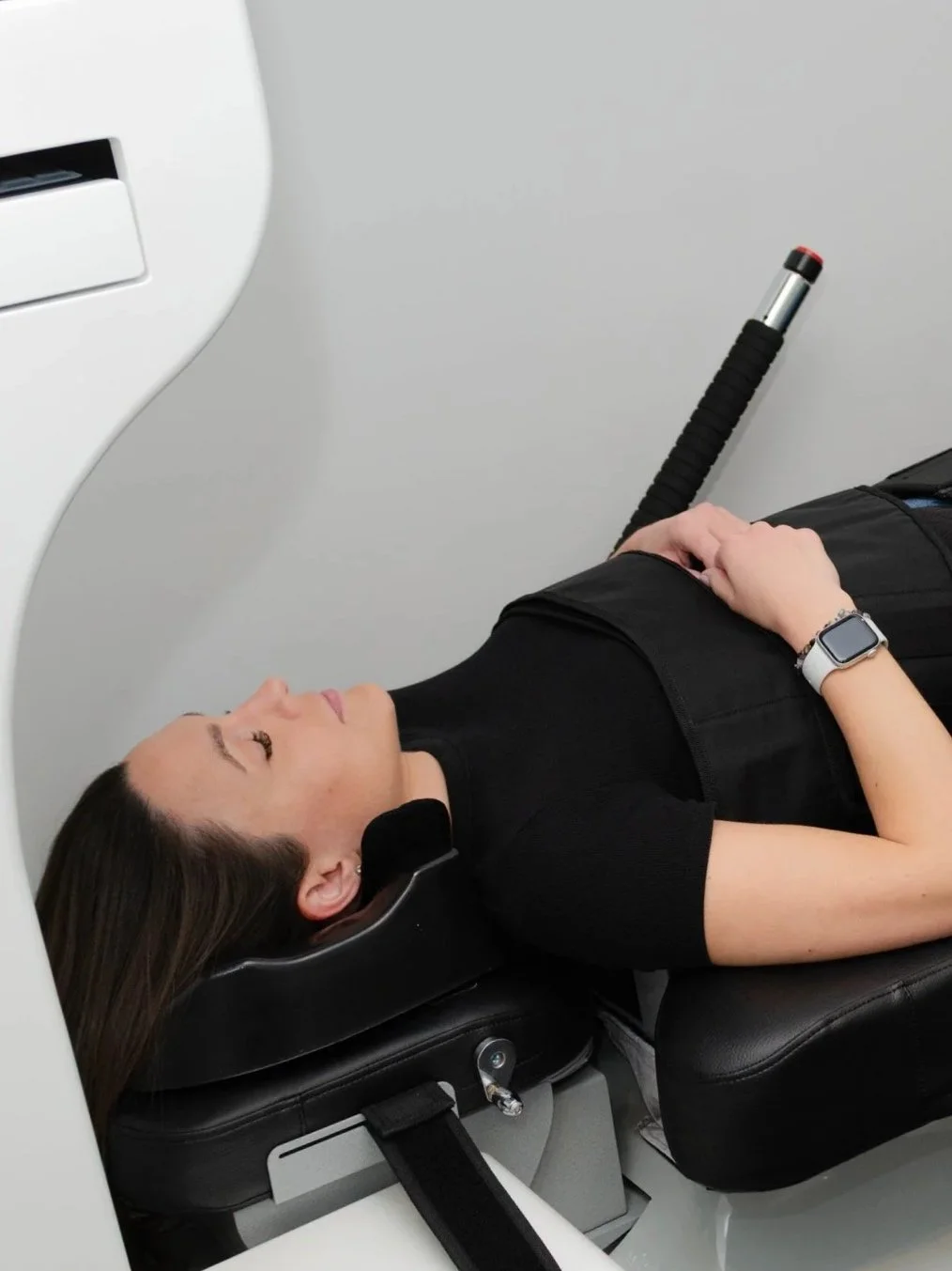 Woman lying down with her head on a cushioned support, preparing for a medical scan, with her hands resting on her stomach, wearing a smartwatch.