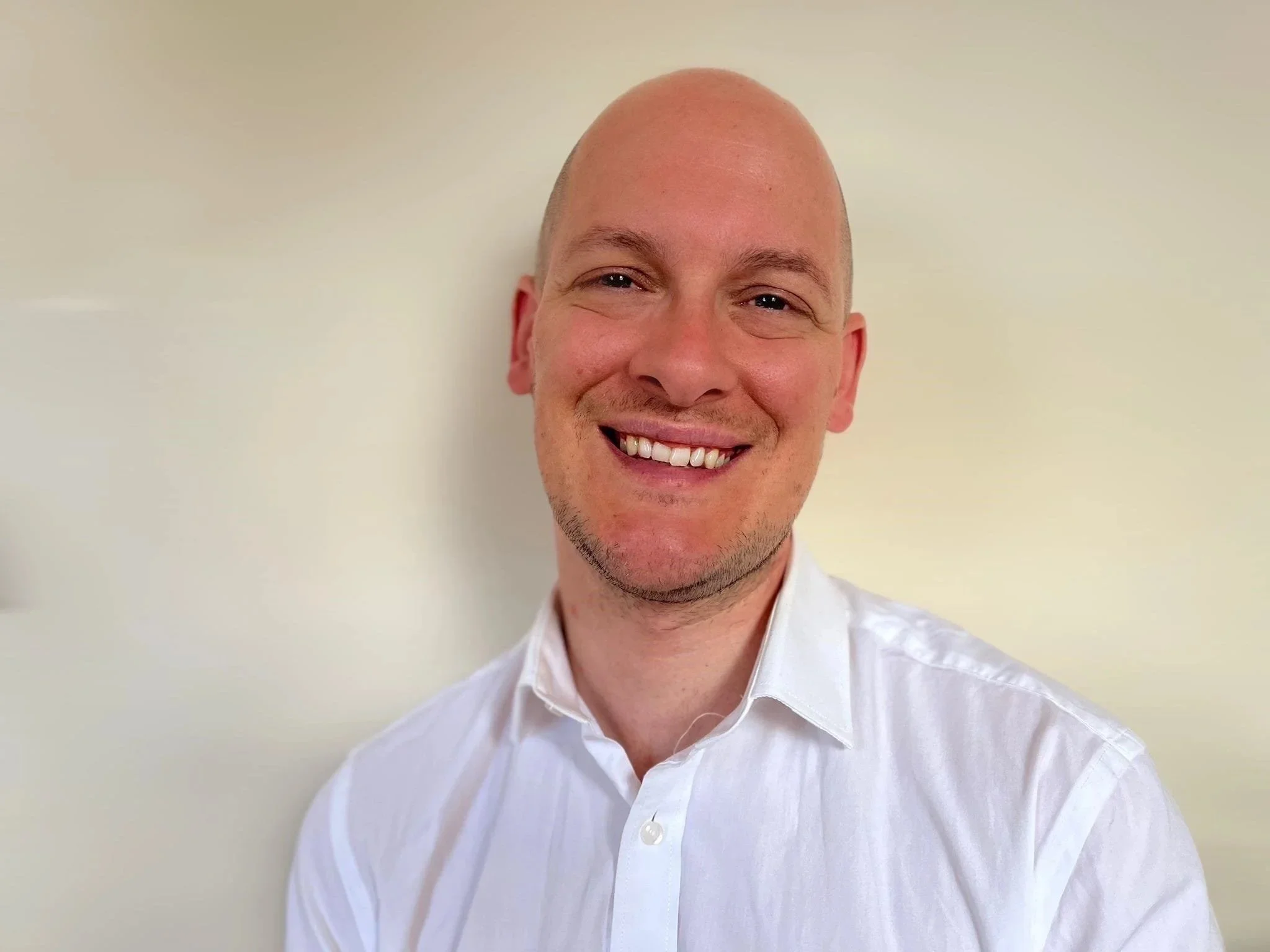 A smiling man with a bald head, wearing a white collared shirt, against a light-colored background.
