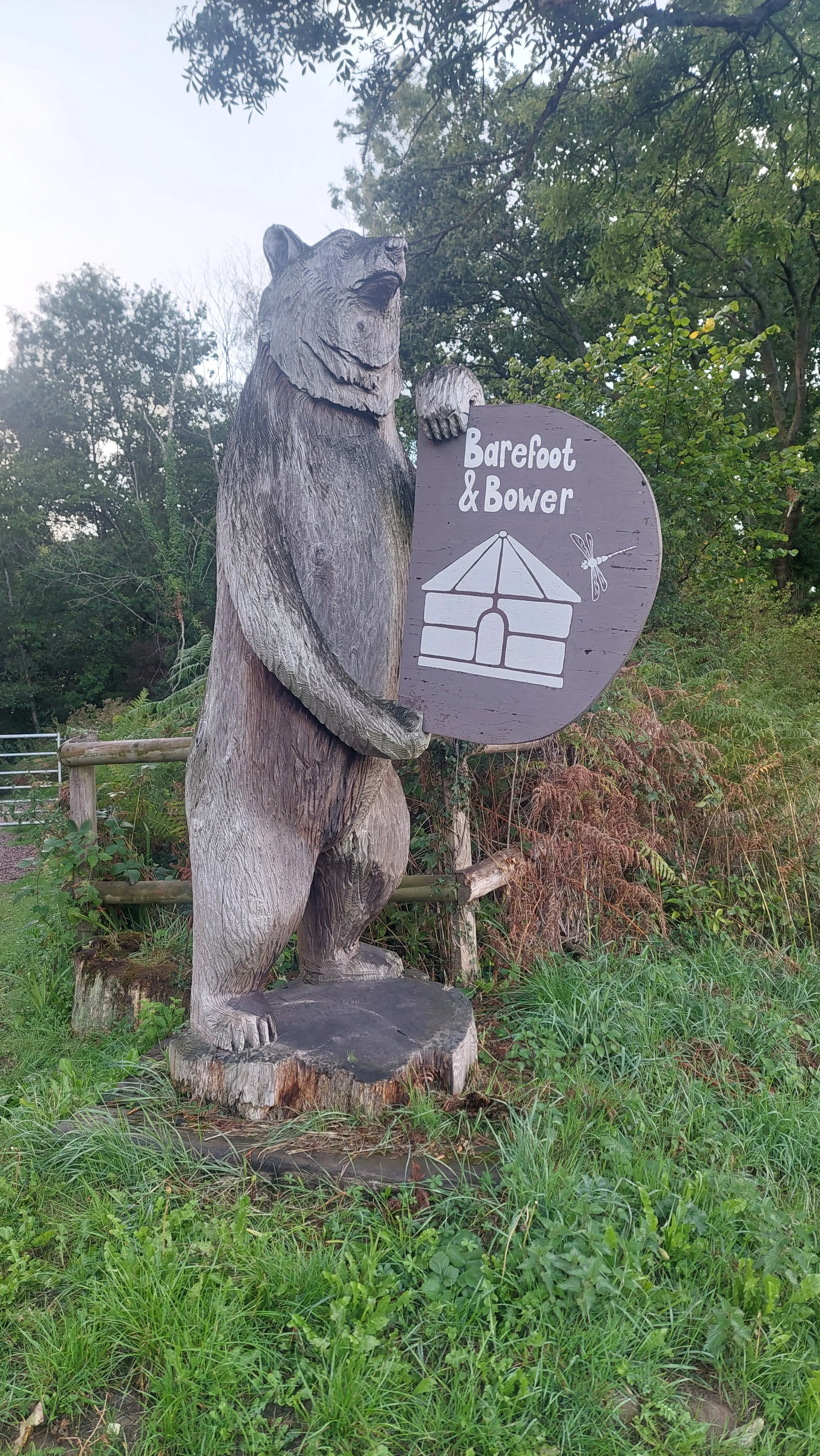 Wooden bear sculpture holding a sign that says 'Barefoot & Bower' with a drawing of a house and a dragonfly, standing on a tree stump in a grassy area.