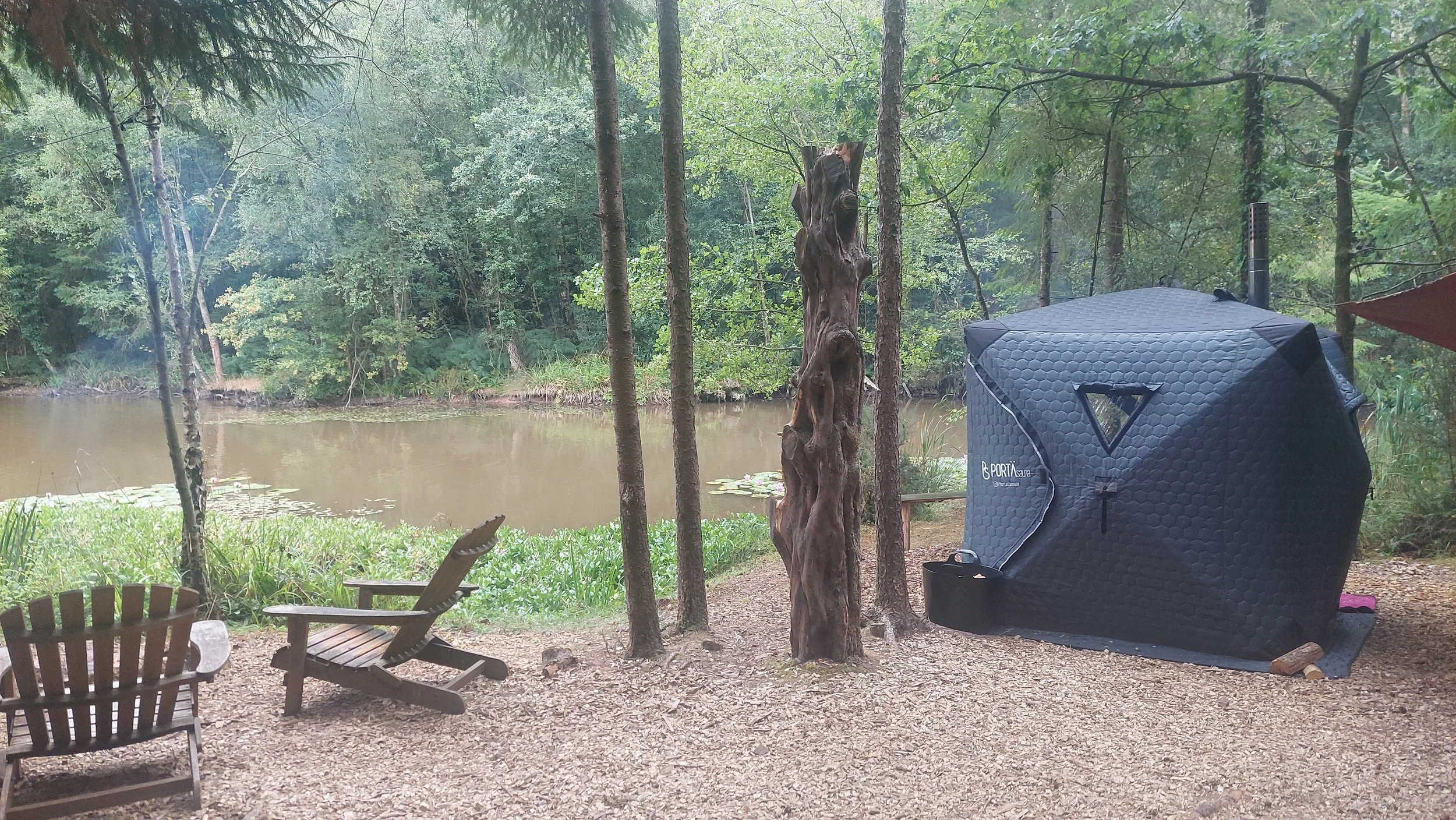 Camping setup by a river with wooden chairs, trees, and a portable sauna in a forested area.