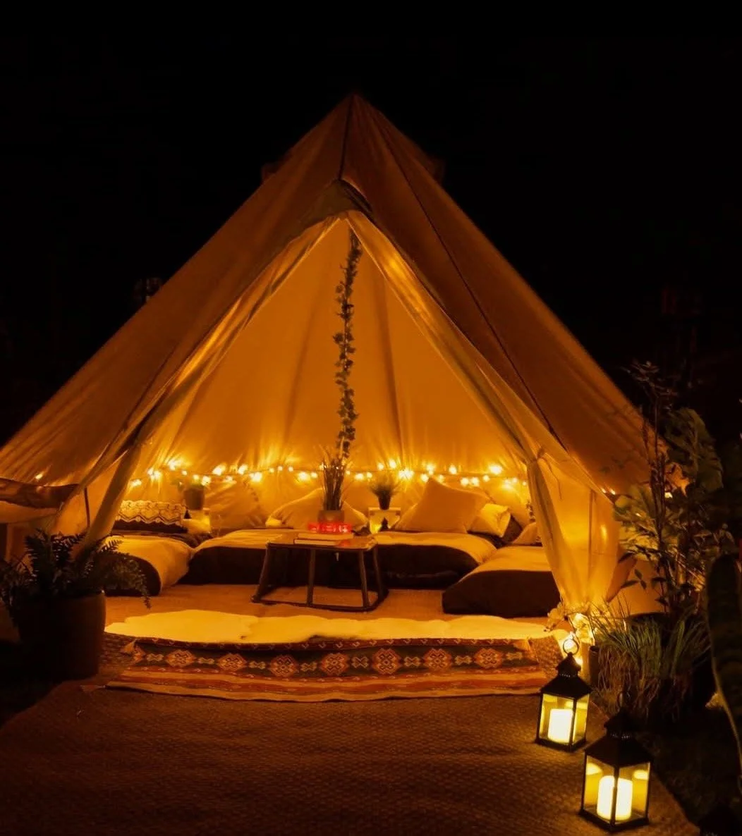 A cozy outdoor tent with warm yellow lighting, decorated with string lights, pillows, and plants, creating an inviting space at night.
