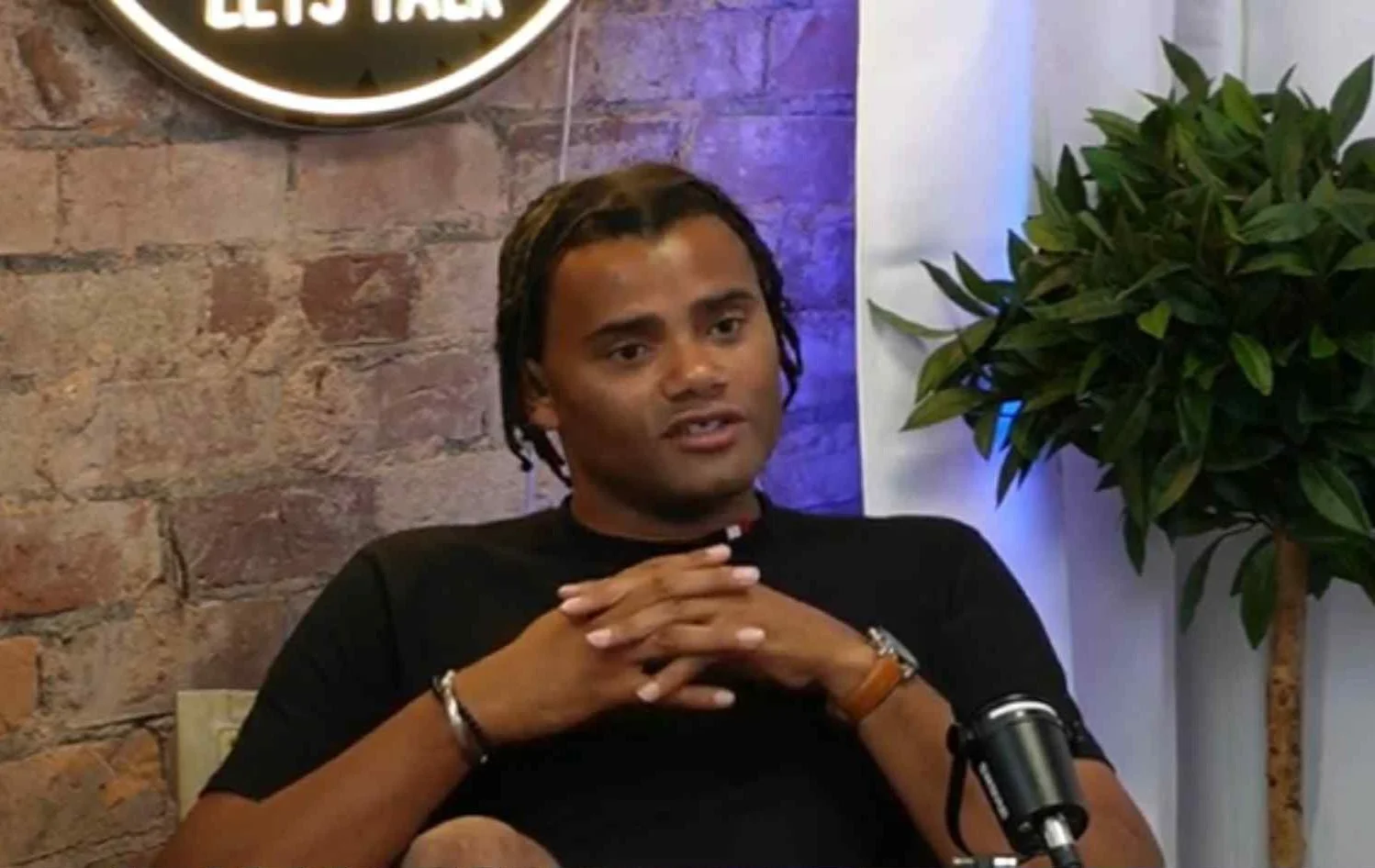A man sitting at a table with hands clasped, speaking or listening, with a brick wall background and a large green plant nearby.