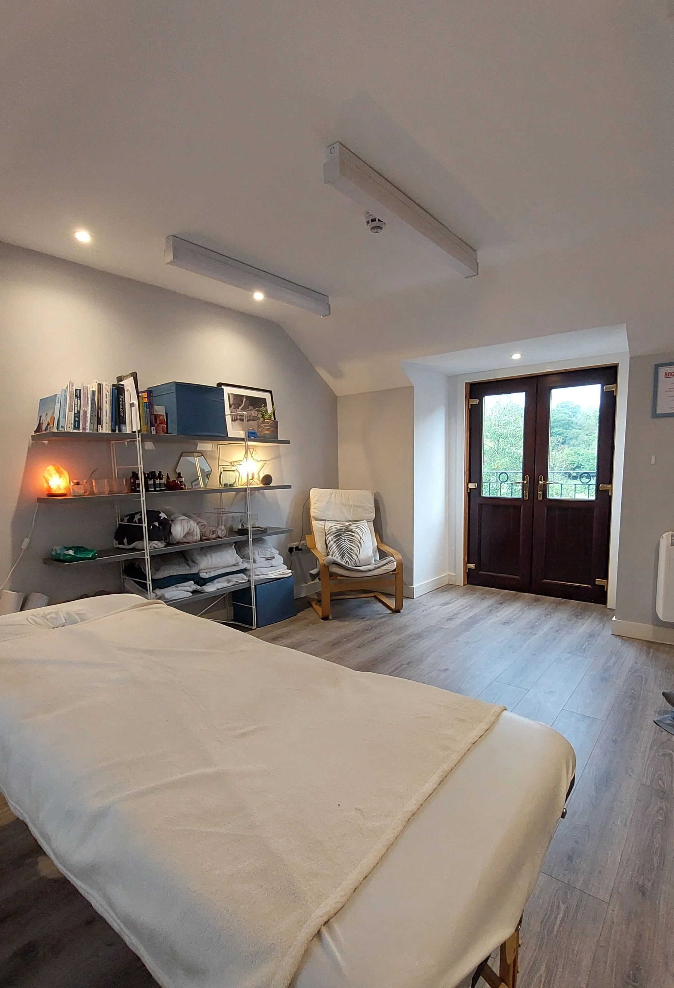 A cozy bedroom with a bed covered in a light-colored blanket, a bookshelf filled with books and decorative items, a wooden chair with a cushion near the window, and a door leading outside. The room has wooden flooring and ceiling lights.