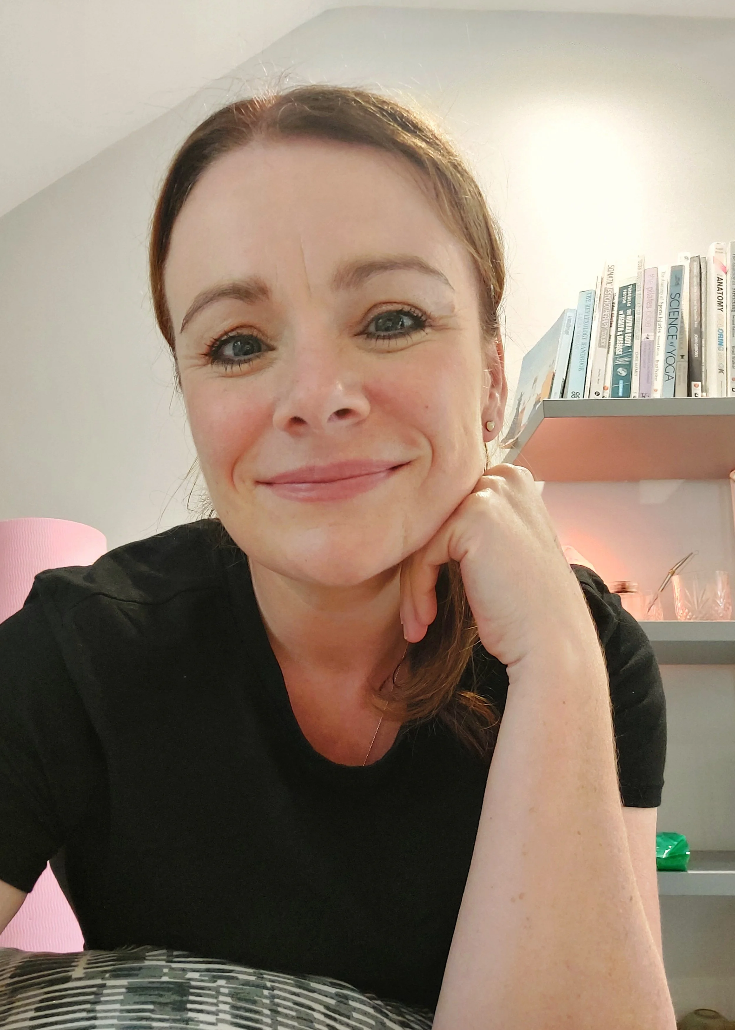 A woman with red hair smiling, resting her chin on her hand, in a room with a bookshelf and pink lamp in the background.