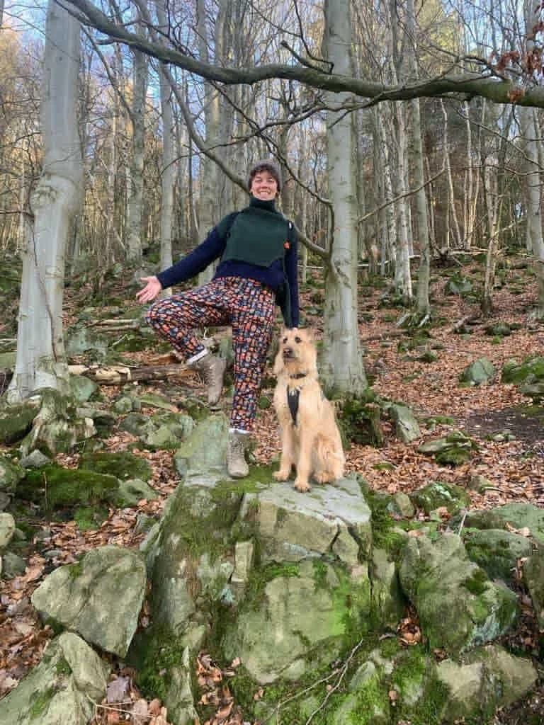 A woman smiling and standing on a large moss-covered rock in a forest, with one foot on a smaller rock, next to a tan dog sitting on the same large rock.