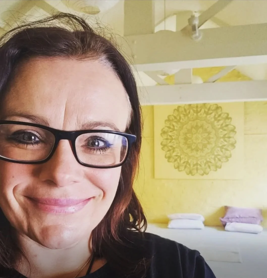 A woman with glasses smiling in a brightly decorated room with yellow walls, a circular patterned artwork, and folded blankets or towels on a shelf in the background.