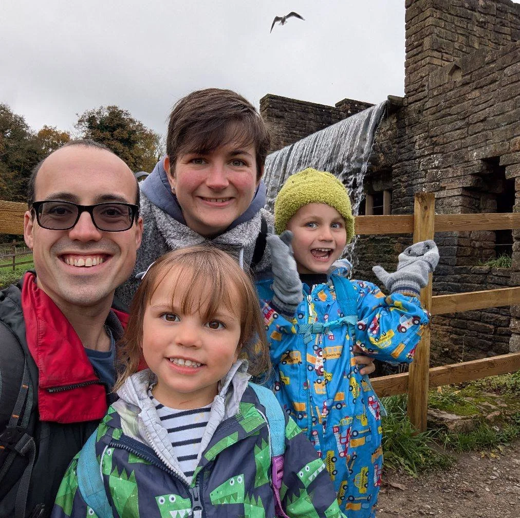 A family of five outdoor during daytime, with a watermill and waterfall in the background, all smiling, wearing rain jackets and gloves.