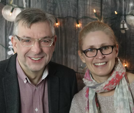 A smiling man and woman wearing glasses, standing indoors with string lights in the background.