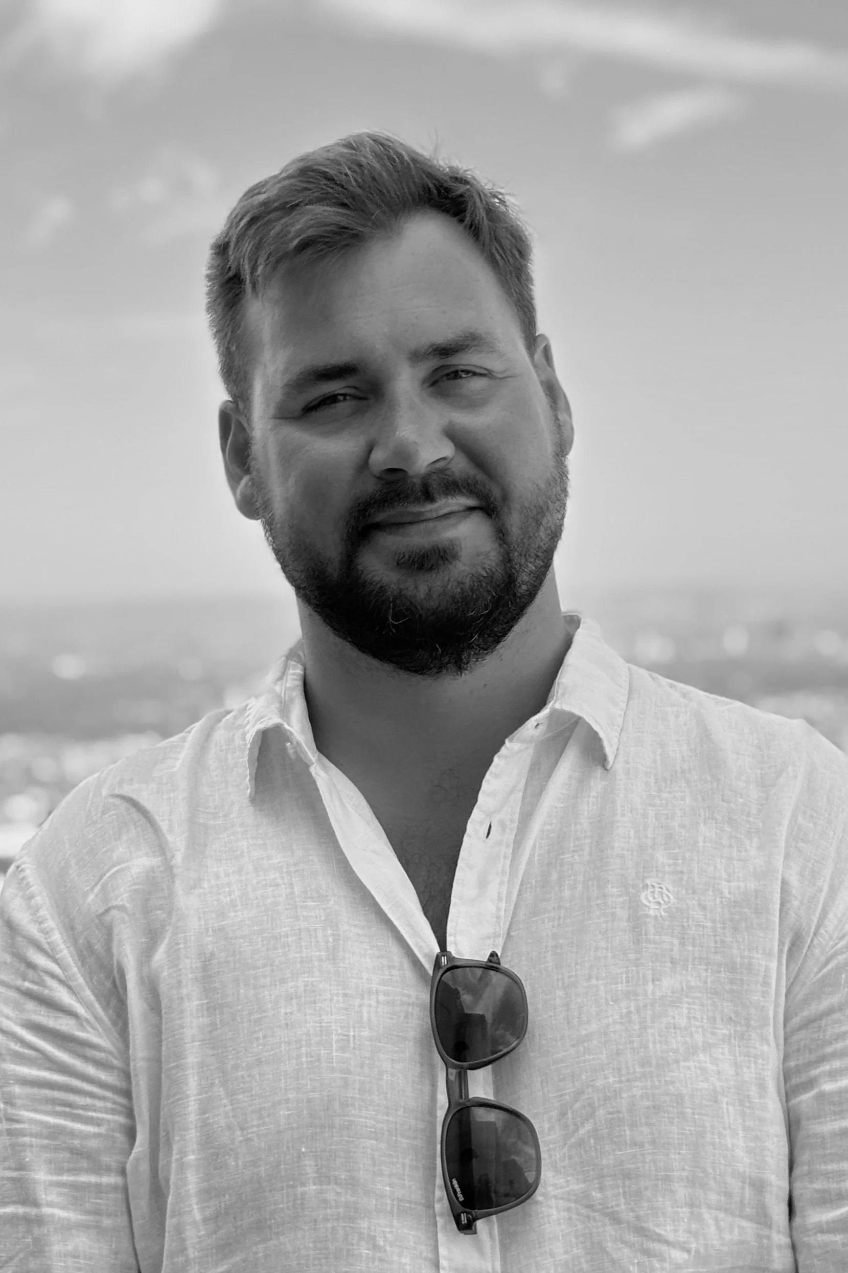 Black and white photo of a man with short, slightly wavy hair, a beard, and mustache, wearing a light-colored button-up shirt with sunglasses hanging from the chest, outdoors with cloudy sky in the background.