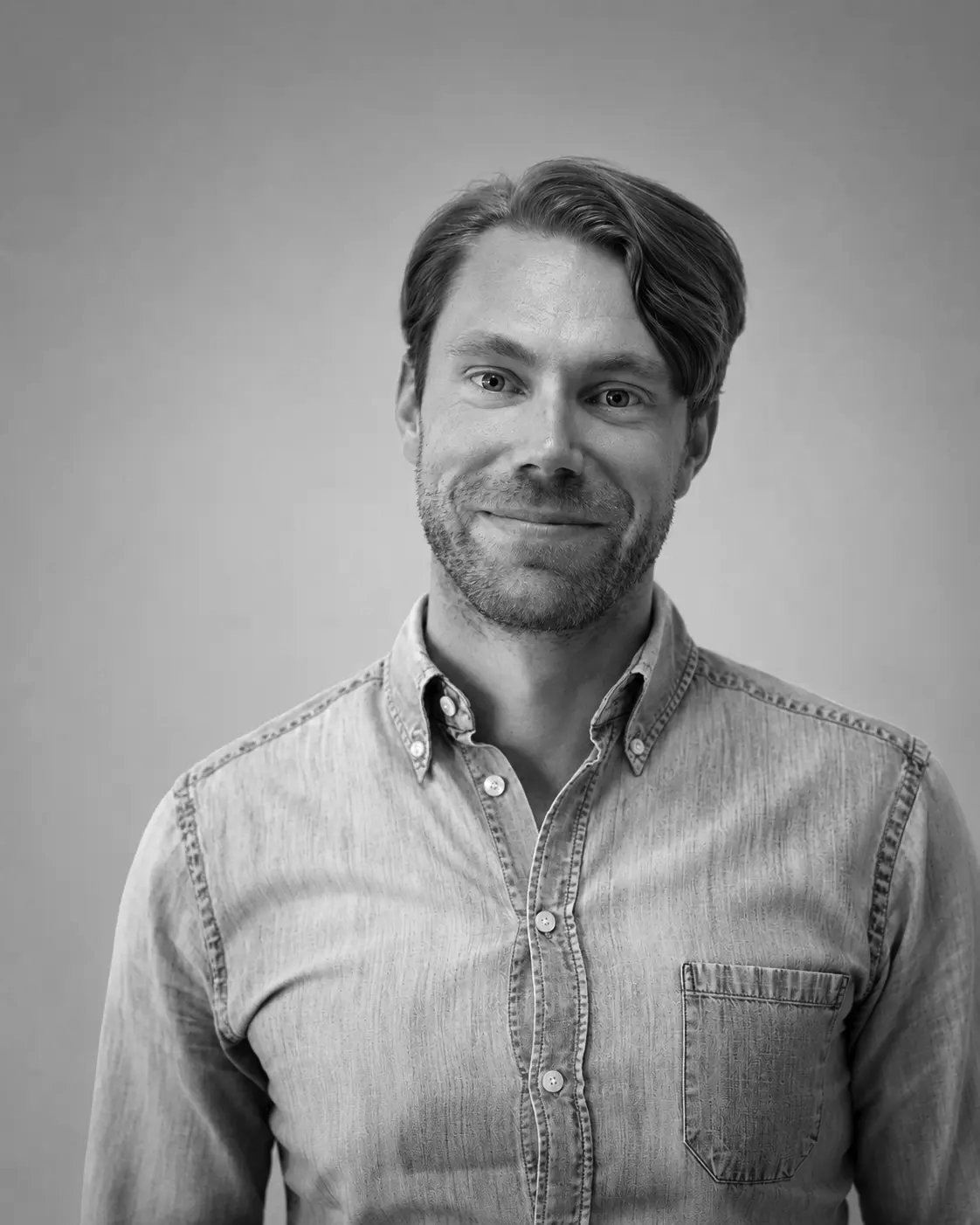 Black and white portrait of a man with light skin, wavy hair, and a beard, smiling slightly. He is wearing a light-colored, button-up denim shirt, standing against a plain background.