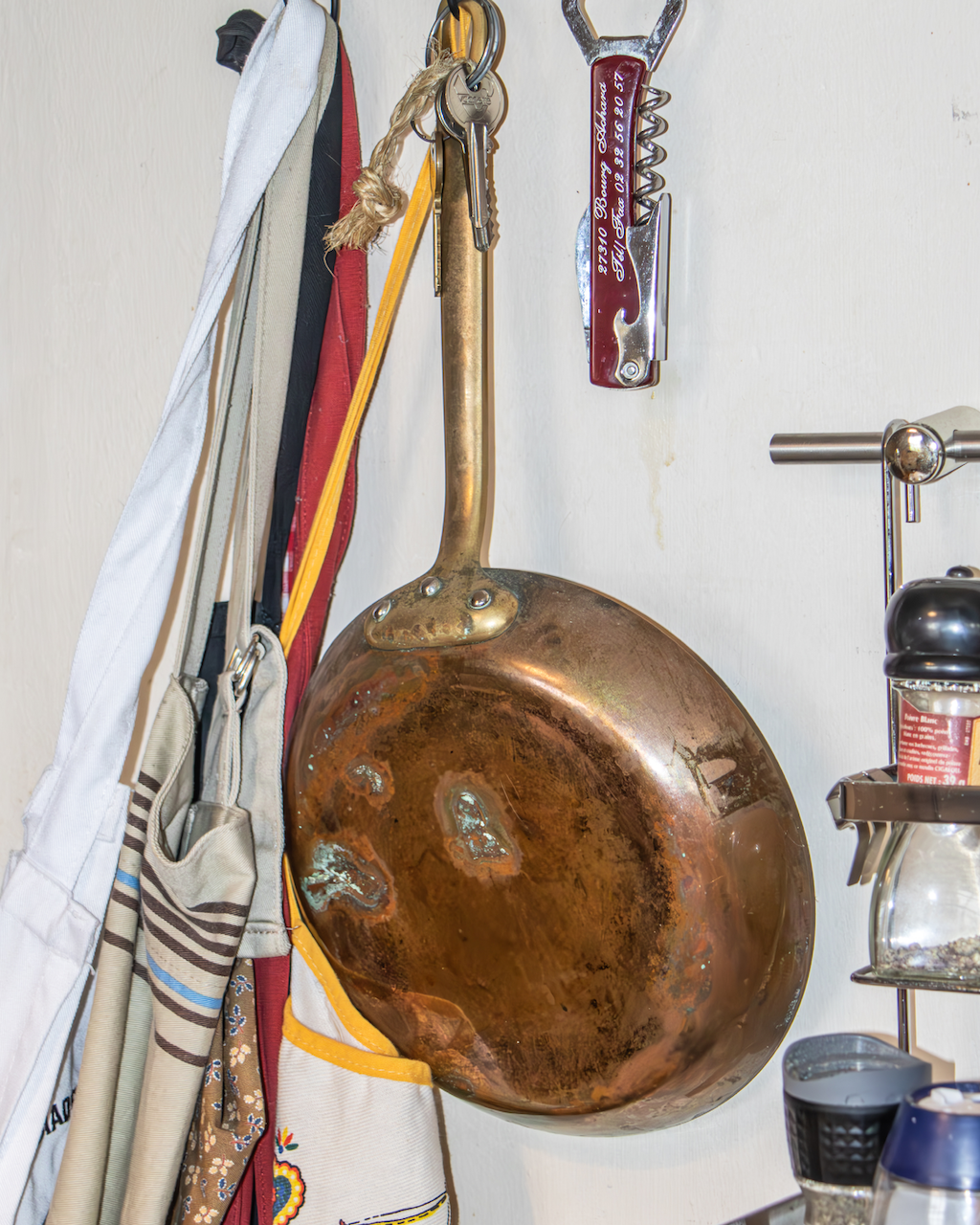 A large, rusty, copper frying pan hanging on a wall beside various hanging kitchen utensils and cloths.
