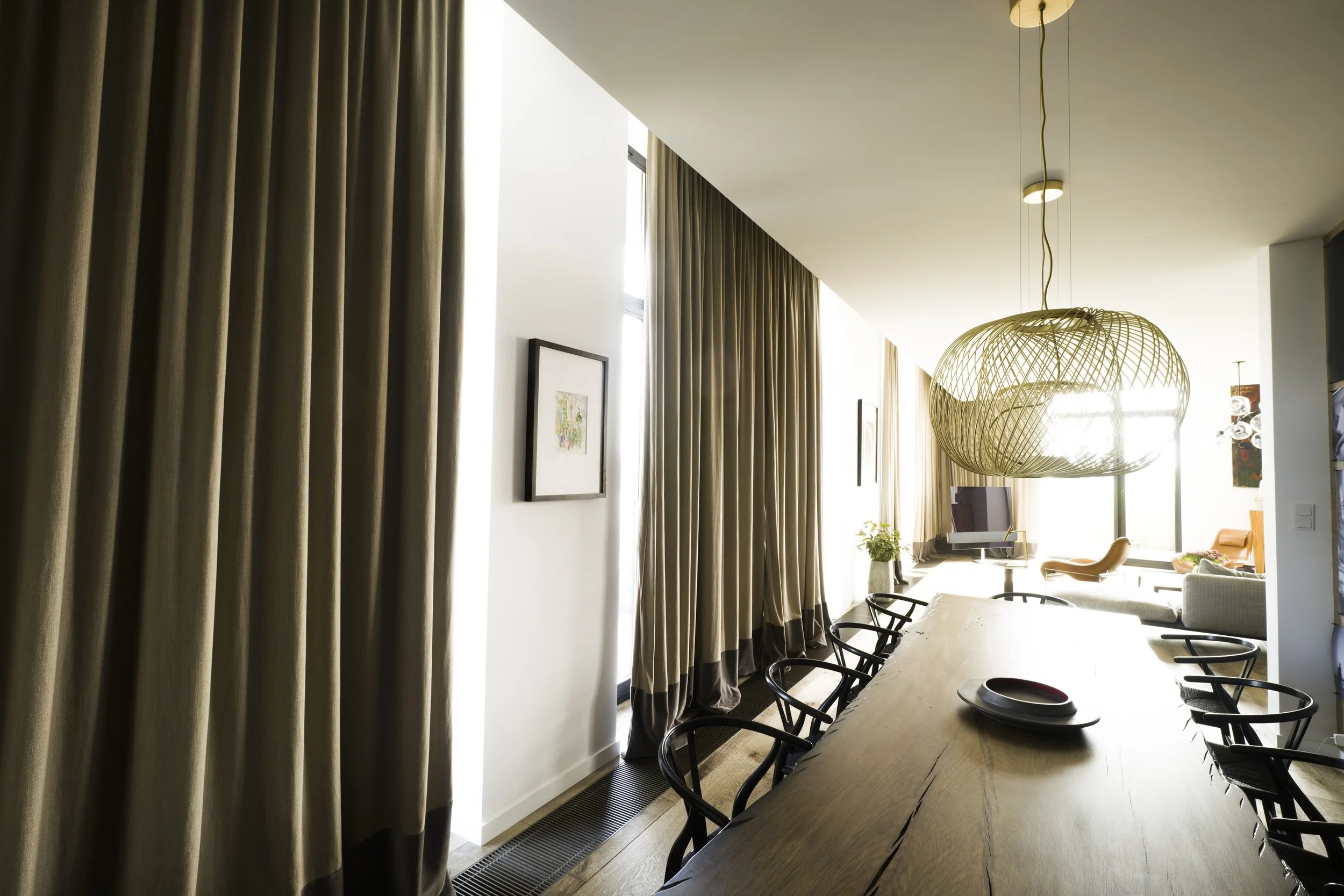 Modern living and dining room with floor-to-ceiling beige curtains, a large oval woven pendant light, a long dining table with black chairs, artwork on white walls, a flat-screen TV, and seating area with chairs and a side table near large windows.