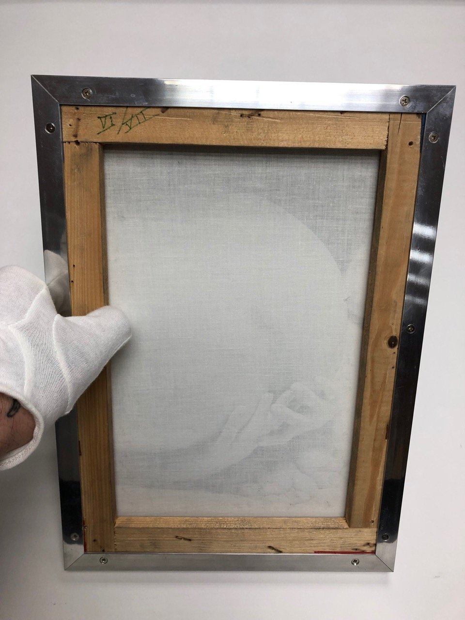 Back of a picture frame with a wooden inner border, metal outer frame, and white backing, held by a person wearing a white glove.
