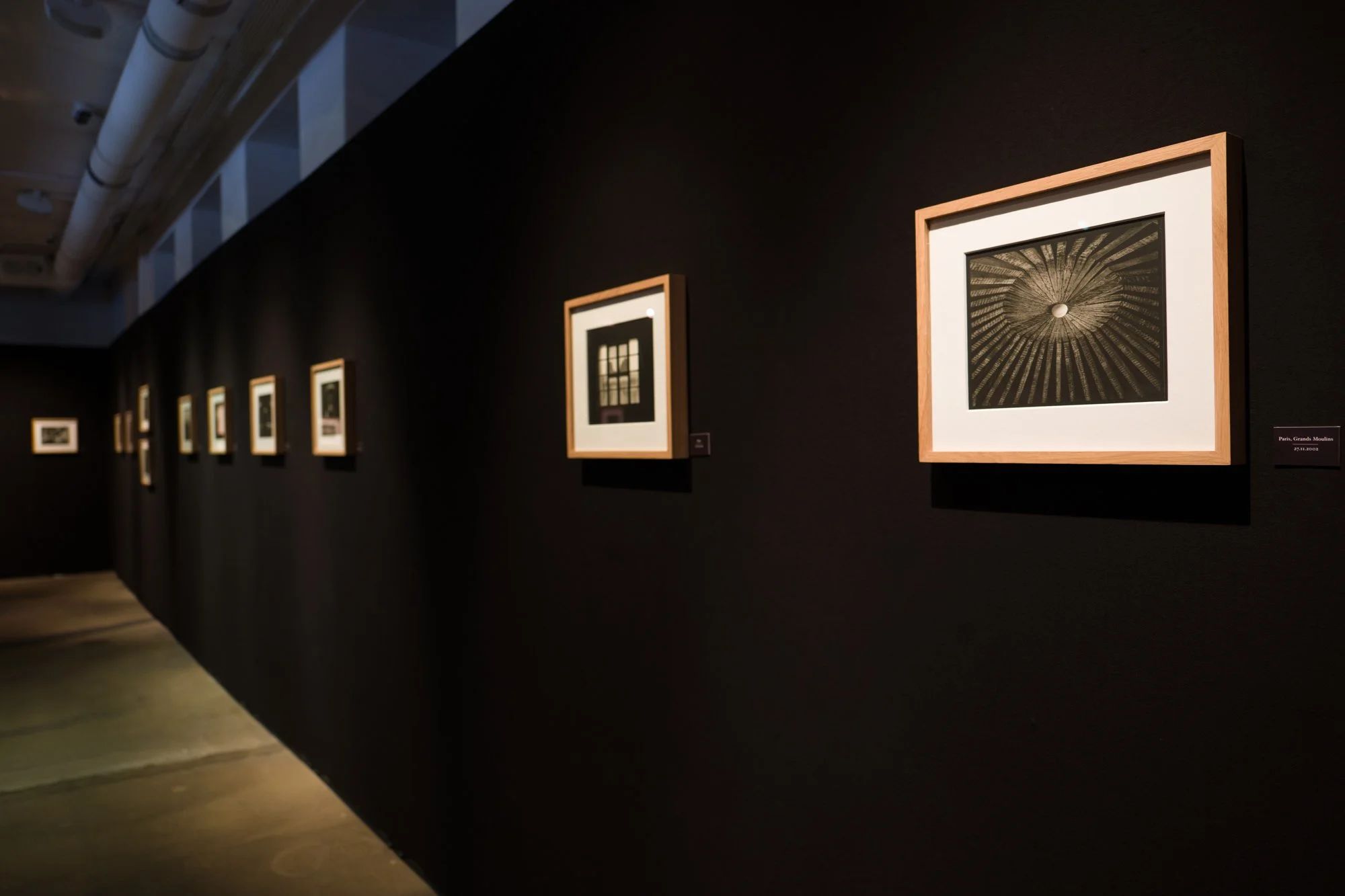 Czarna ściana galerii z oprawionymi w drewniane ramy fotografiami Bogdana Konopki z cyklu Lekcje Ciemności.