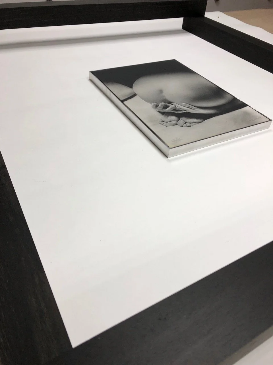 A black and white photograph of a person's hand holding a rounded object, possibly a stone, against their midsection, displayed on a white surface.