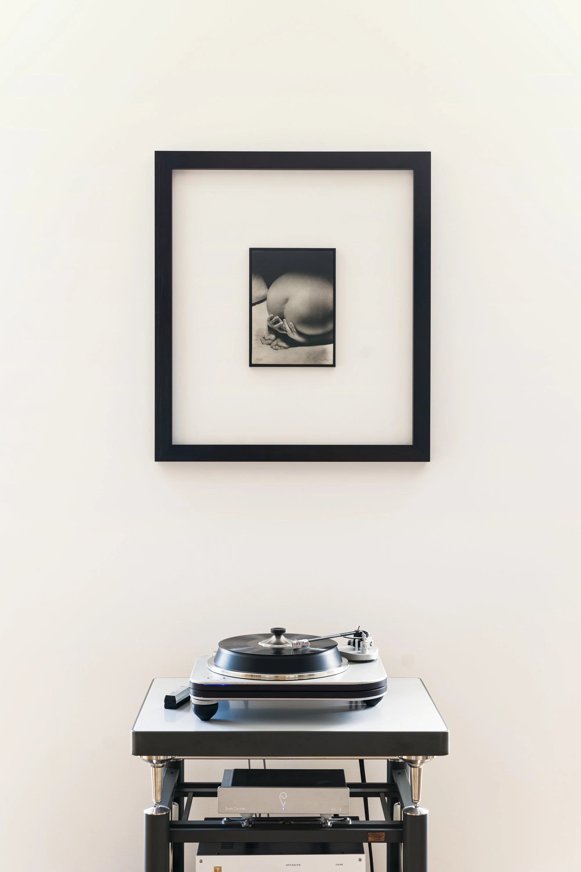 A turntable on a black stand, with a framed monochrome artwork of a person's face and hand on the wall behind it.
