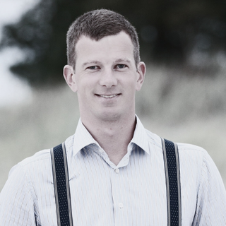 A man with short dark hair wearing a light-colored shirt with suspenders stands outdoors with blurred greenery in the background.
