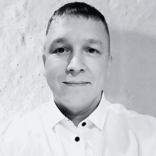 Black and white photo of a man with short hair, wearing a collared shirt, smiling slightly in front of a plain wall.