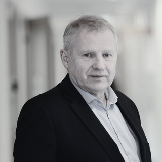 A black-and-white photo of a middle-aged man with short light hair, wearing a dark blazer over a light shirt, standing indoors with a blurred background.