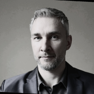 Portrait of a man with short gray hair and beard, wearing a dark blazer and shirt, against a plain background.
