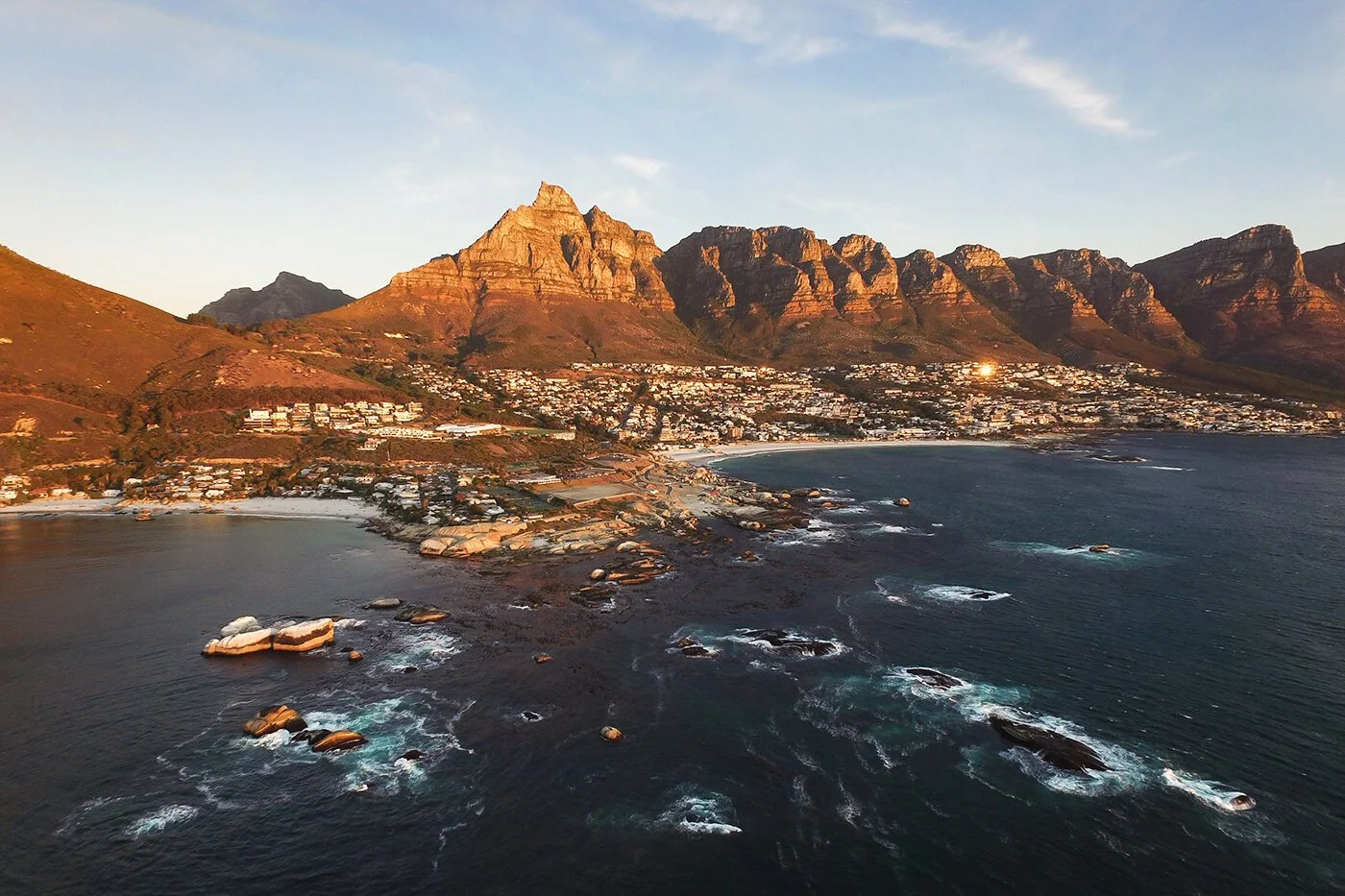 Aerial view of Cape Town with mountains in the background, rocky shoreline, and ocean waves.