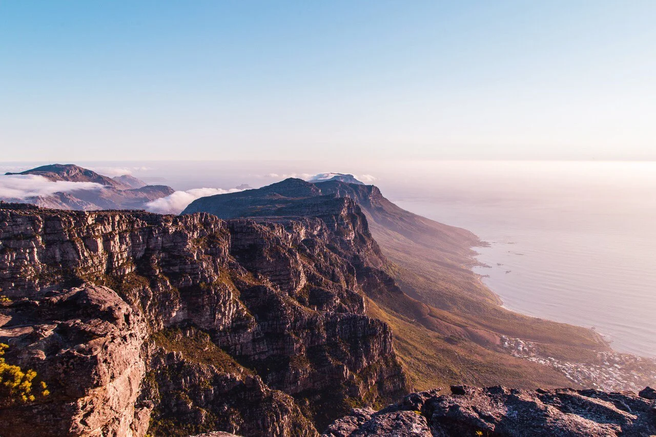 Scenic view of Table Mountain in Cape Town, South Africa, with rugged cliffs, ocean, and misty clouds at sunrise.