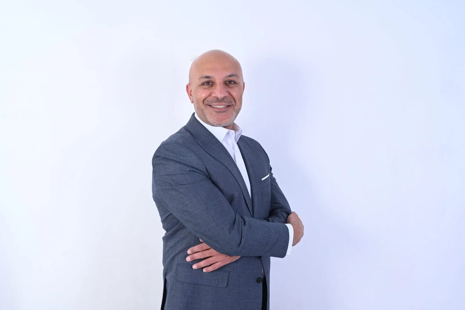 Headshot of a bald man in a navy blazer, white shirt, and light-colored tie, smiling against a plain light gray background.