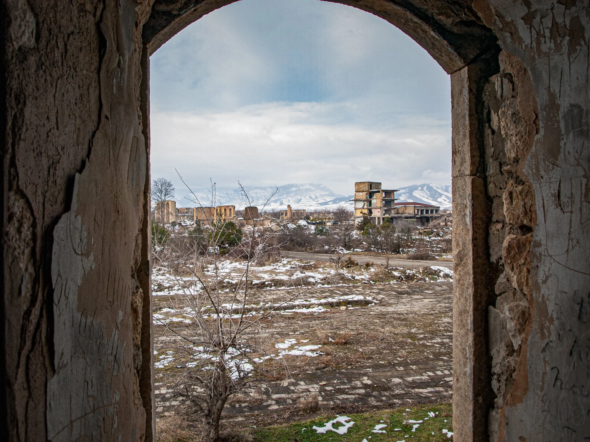 View of abandoned buildings and a snow-covered landscape through a stone archway with weathered walls.